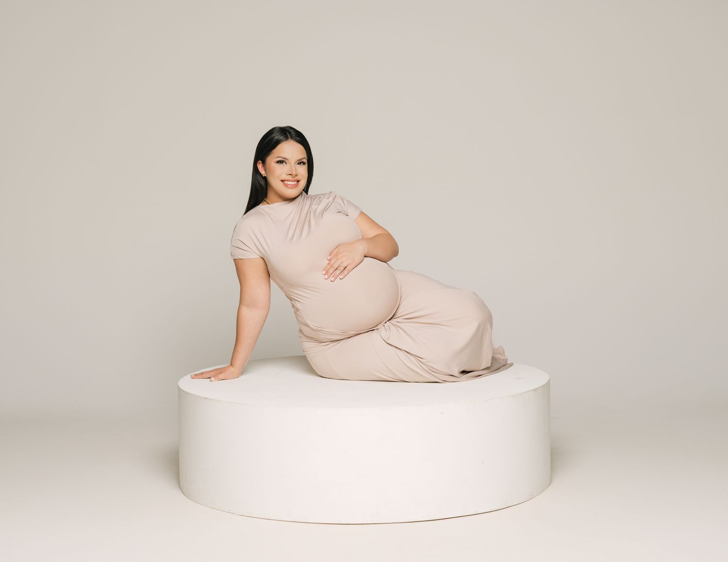 Modern maternity portraits of person in beige dress posing on white circular platform.