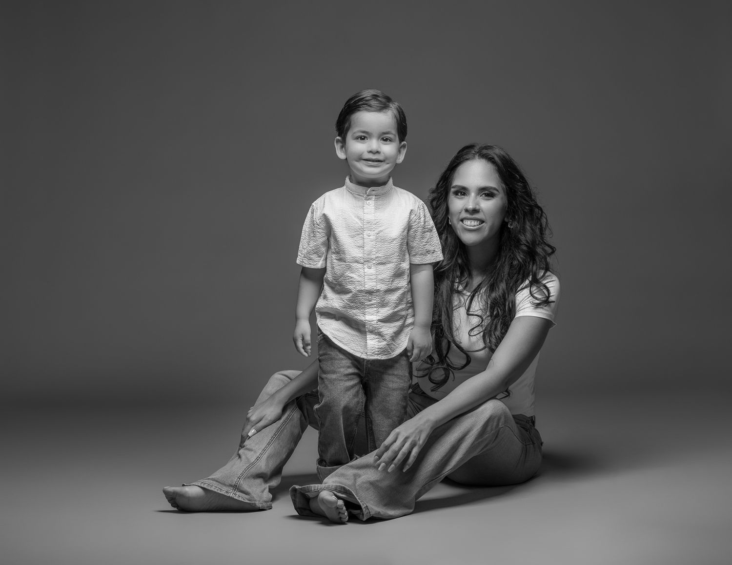 Black and white studio portrait of parent and child sitting together on grey background.