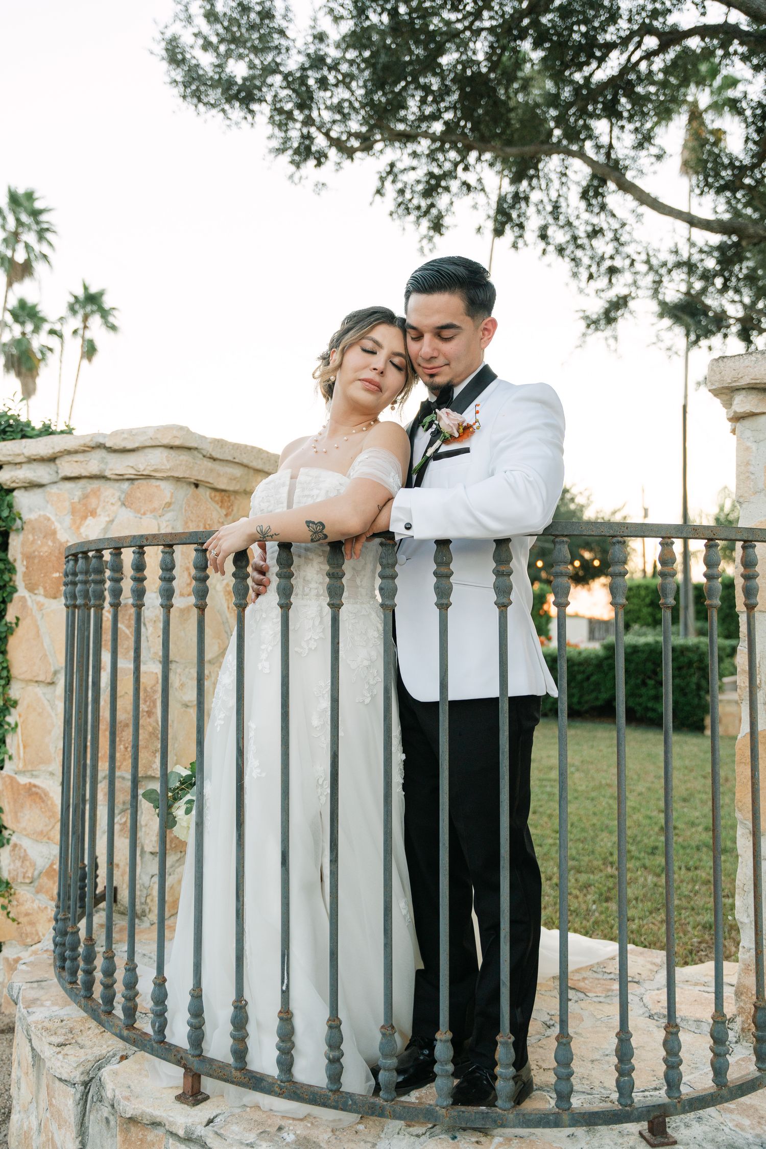 Wedding couple poses romantically along decorative stone balustrade in garden setting.