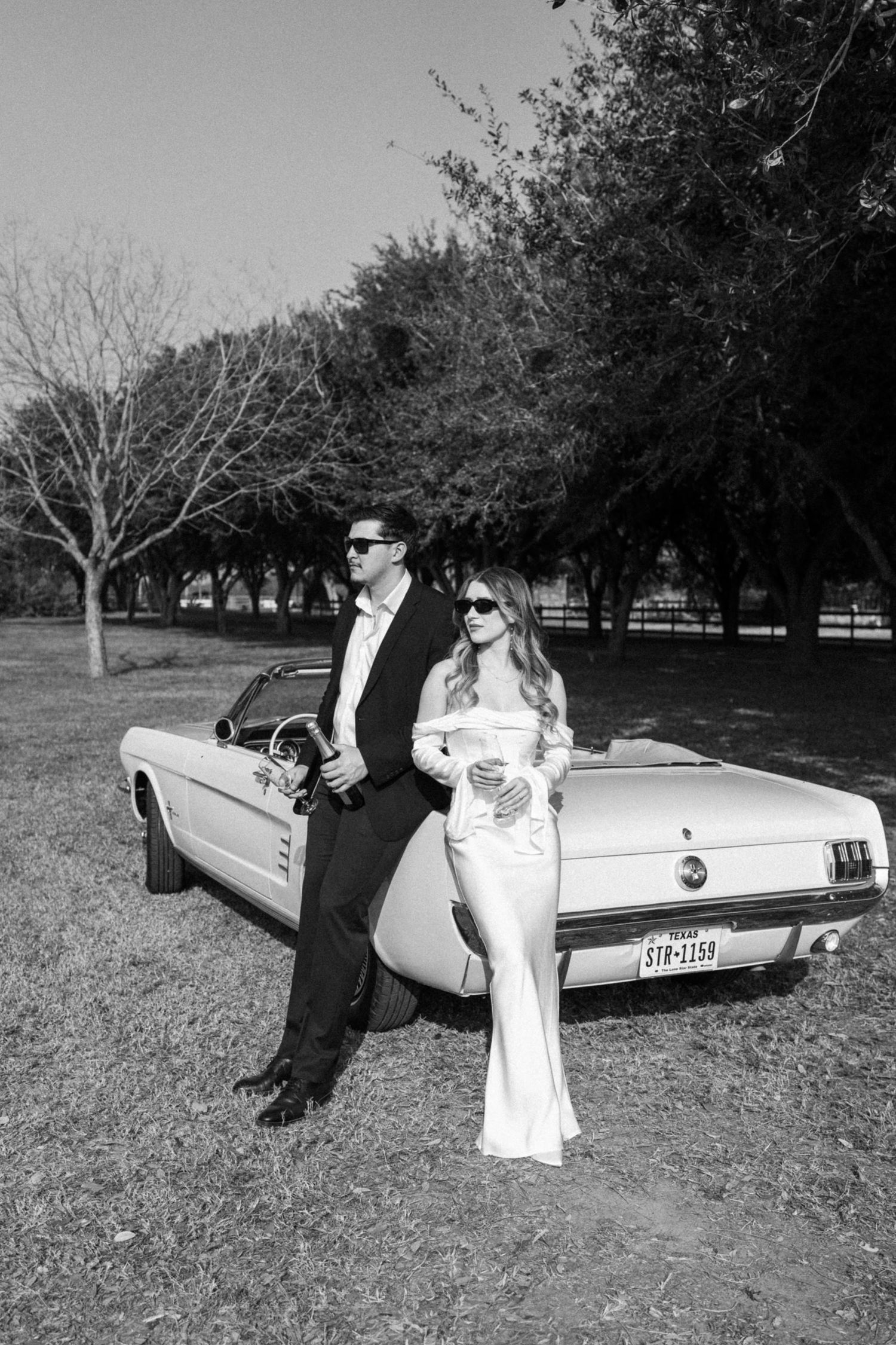 A couple in vintage-inspired attire poses beside a classic Ford Mustang convertible in a park setting.