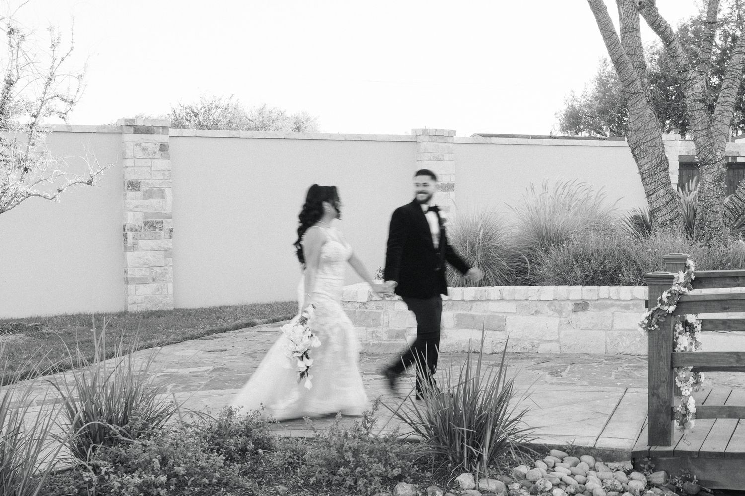 Black and white photo of a bride and groom walking hand in hand through a garden at their outdoor wedding venue.