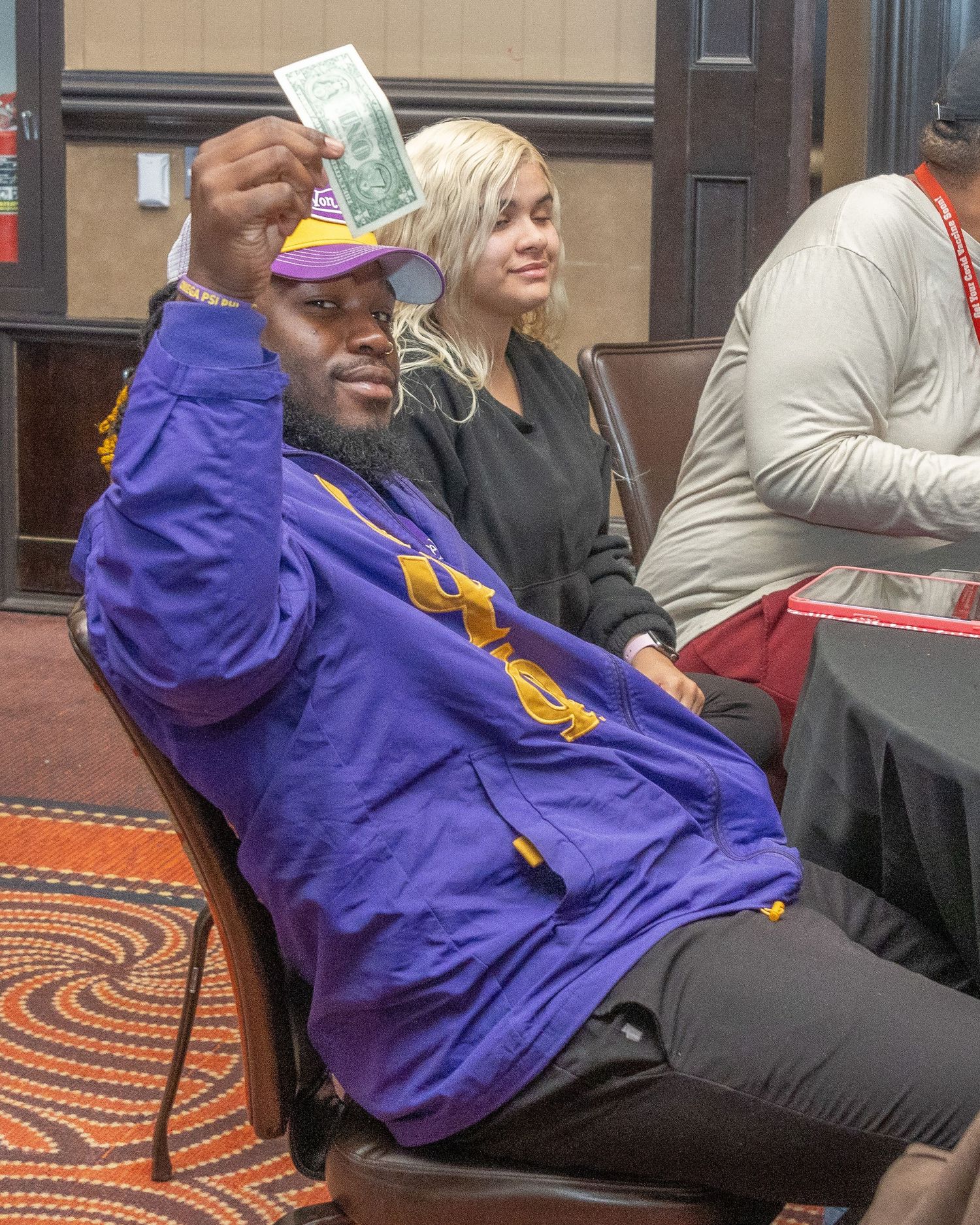 Person in purple jacket holds up money while sitting in conference setting.