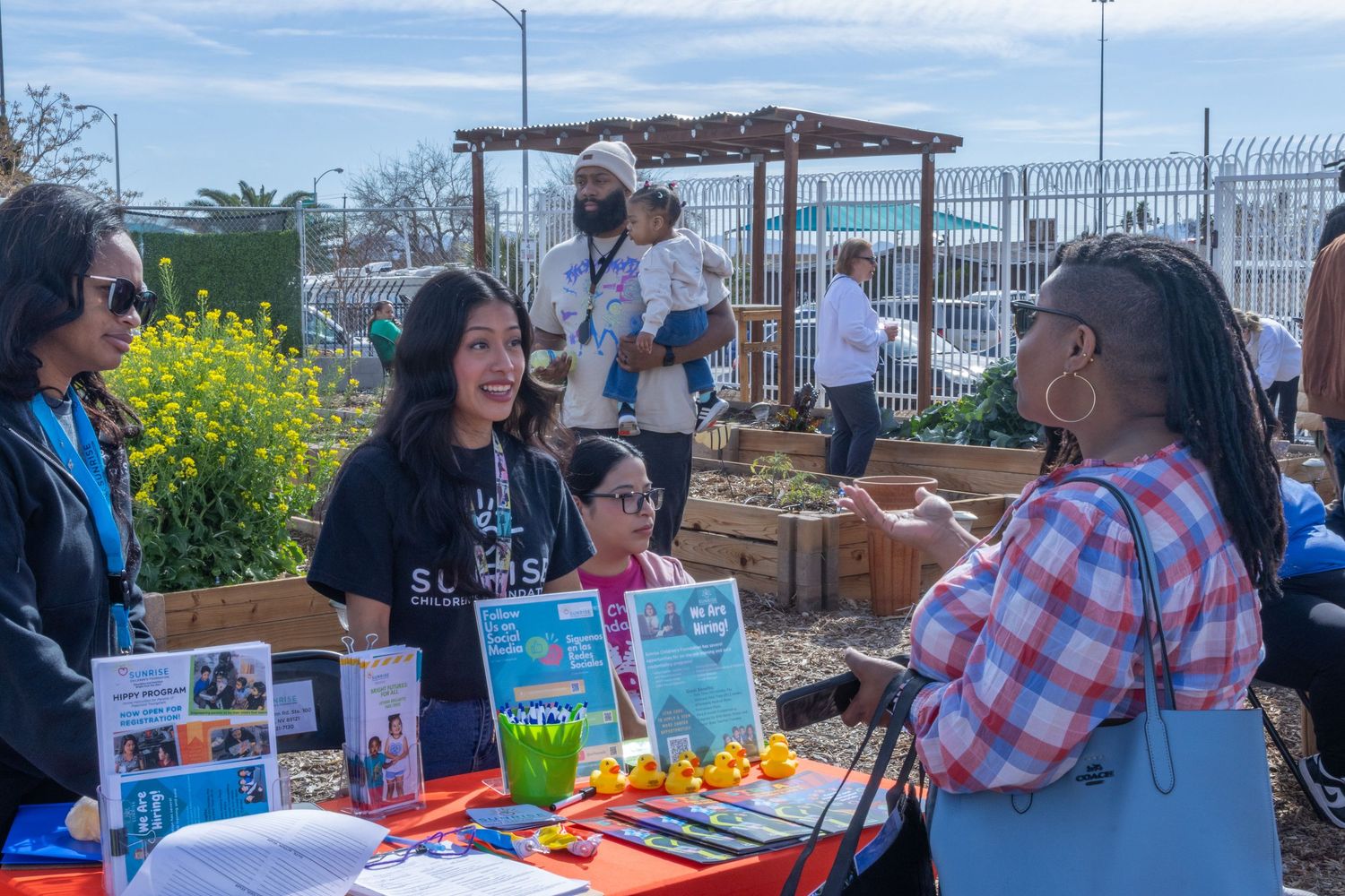 People gather at outdoor tables for a community meal event under blue skies with gardens visible in the background.