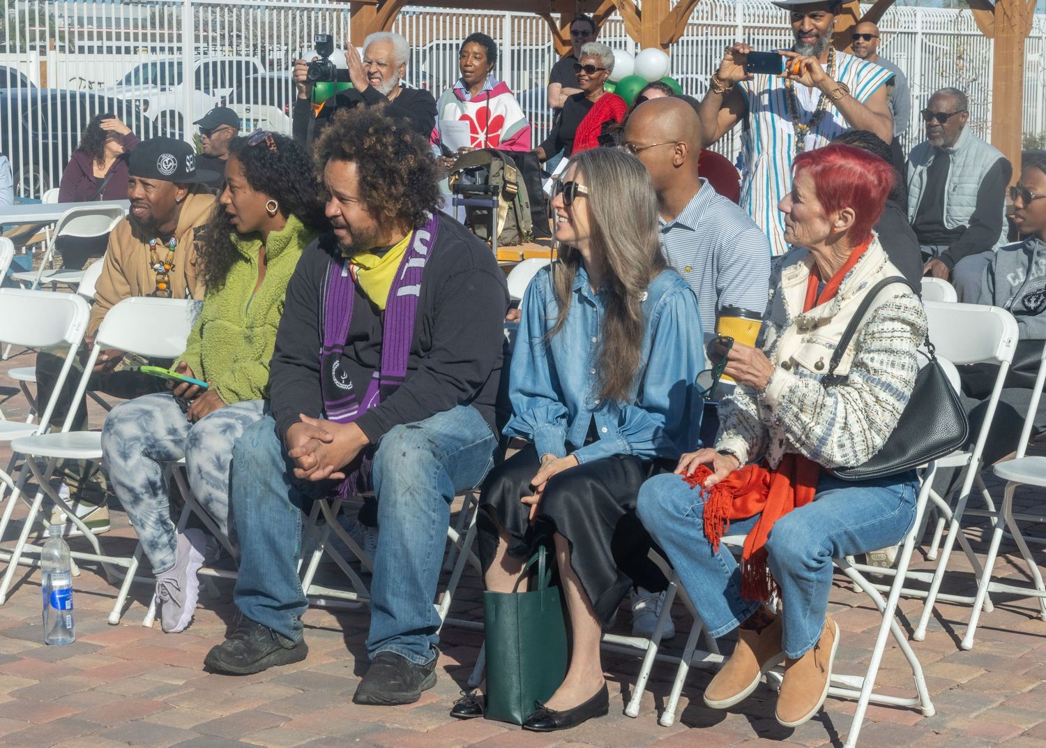 Crowded photo shows seated audience members at a community event with white folding chairs on a wooden floor.