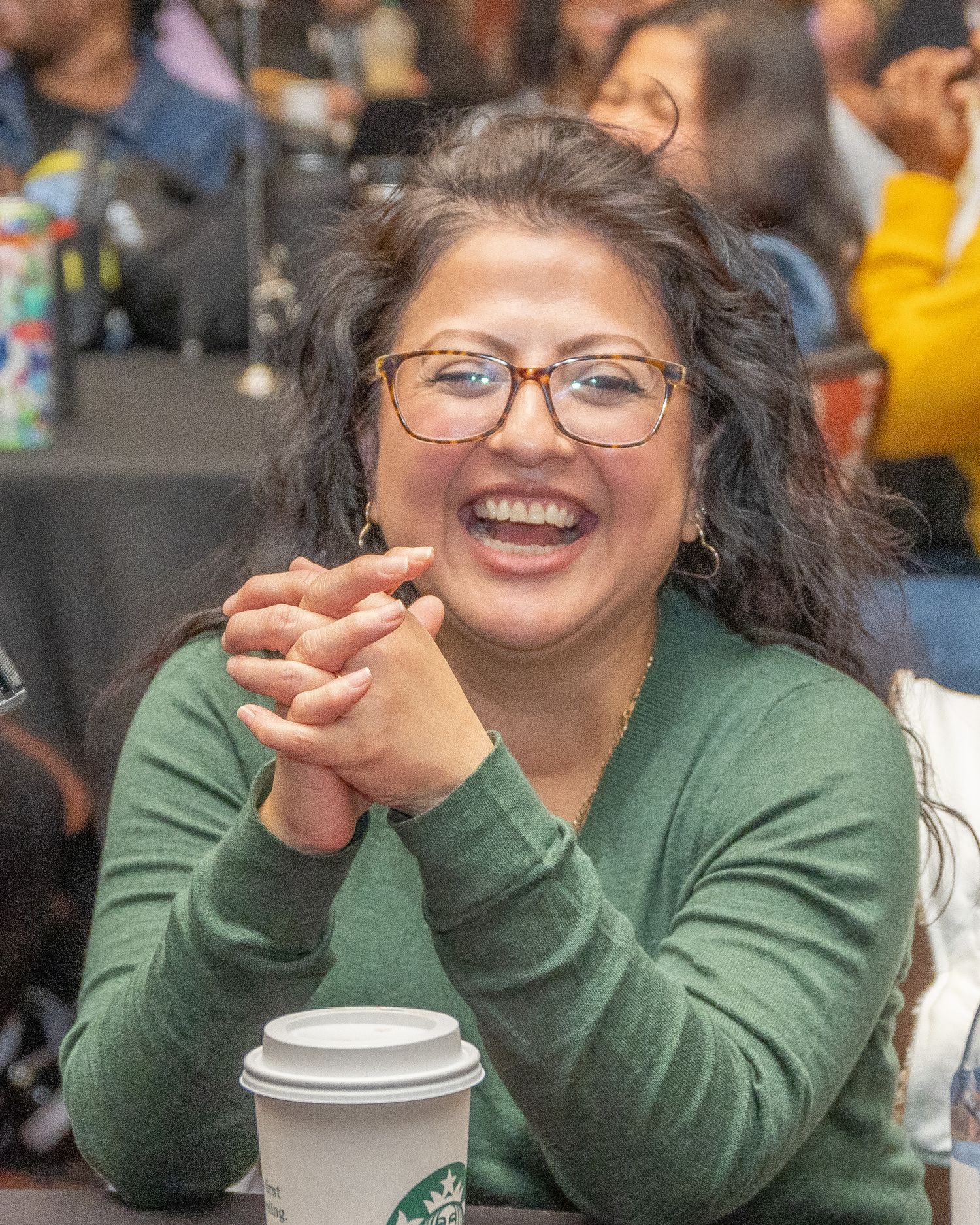 Person wearing glasses and green sweater laughs joyfully while holding a coffee cup at a social gathering.
