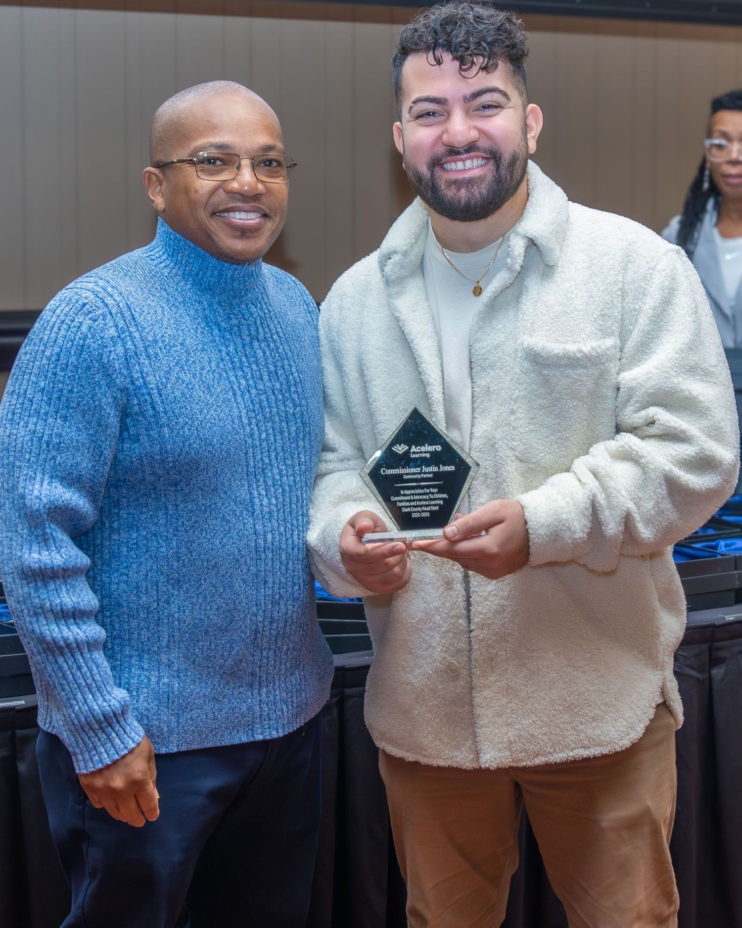 Two people pose together while holding an award plaque at an indoor ceremony event.