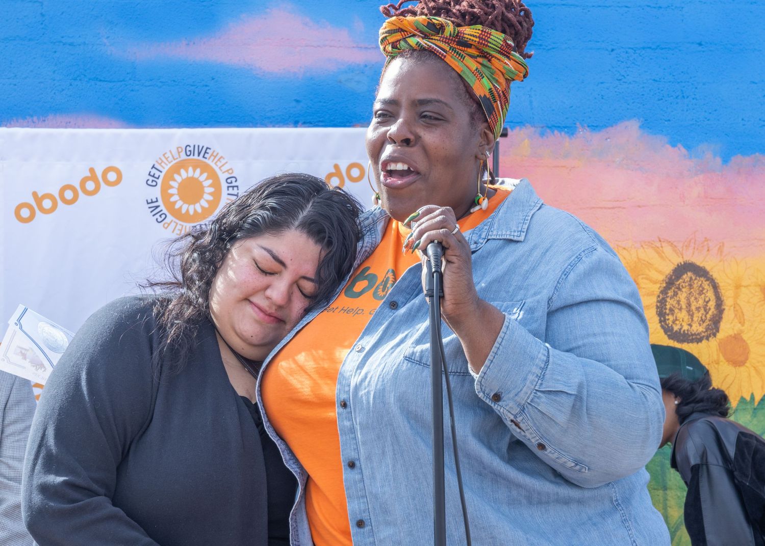 Two people share a joyful moment at an outdoor event against a backdrop of orange and blue artwork.