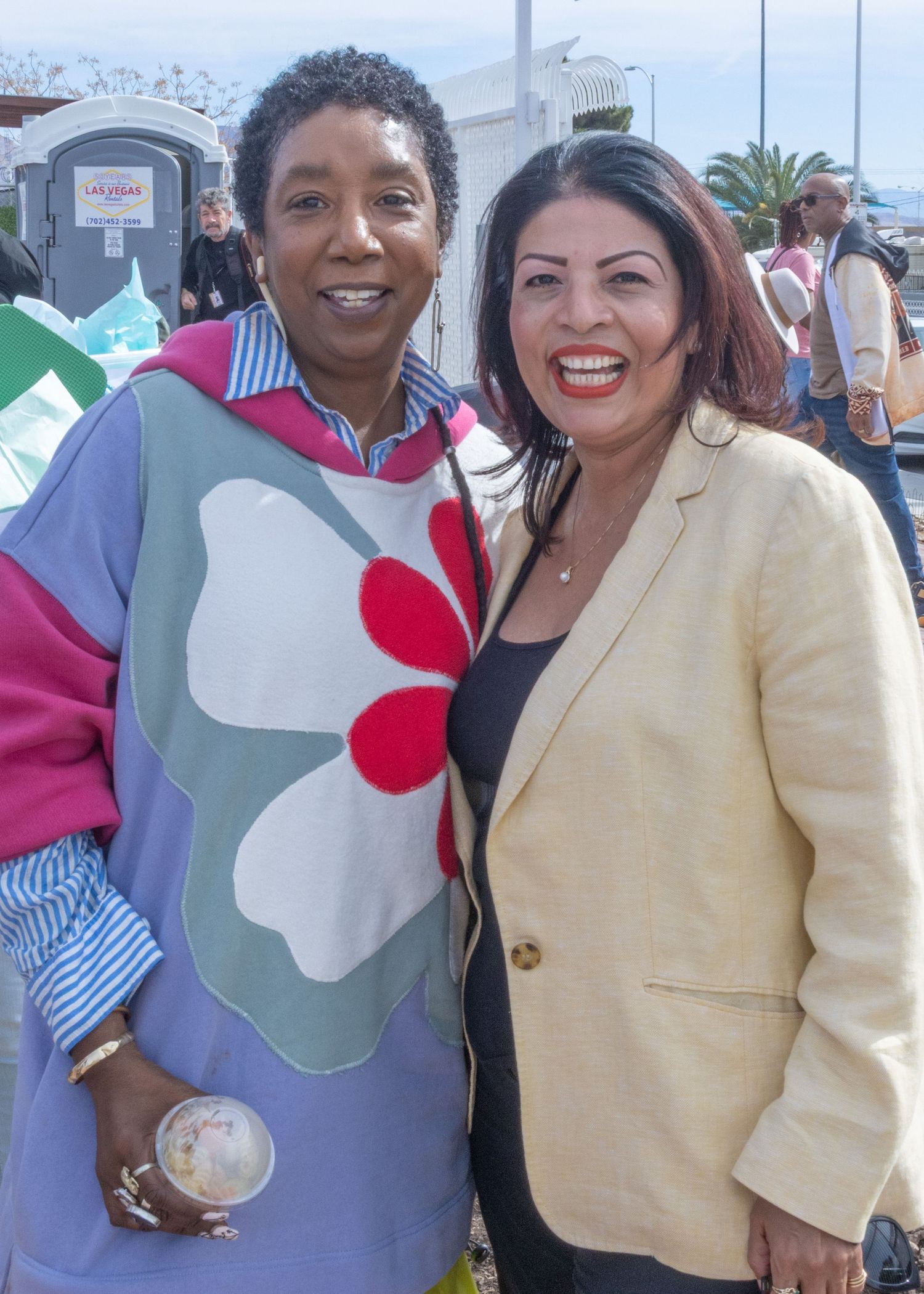 Two smiling individuals pose together outdoors, one wearing a colorful floral sweater and another in a beige blazer.