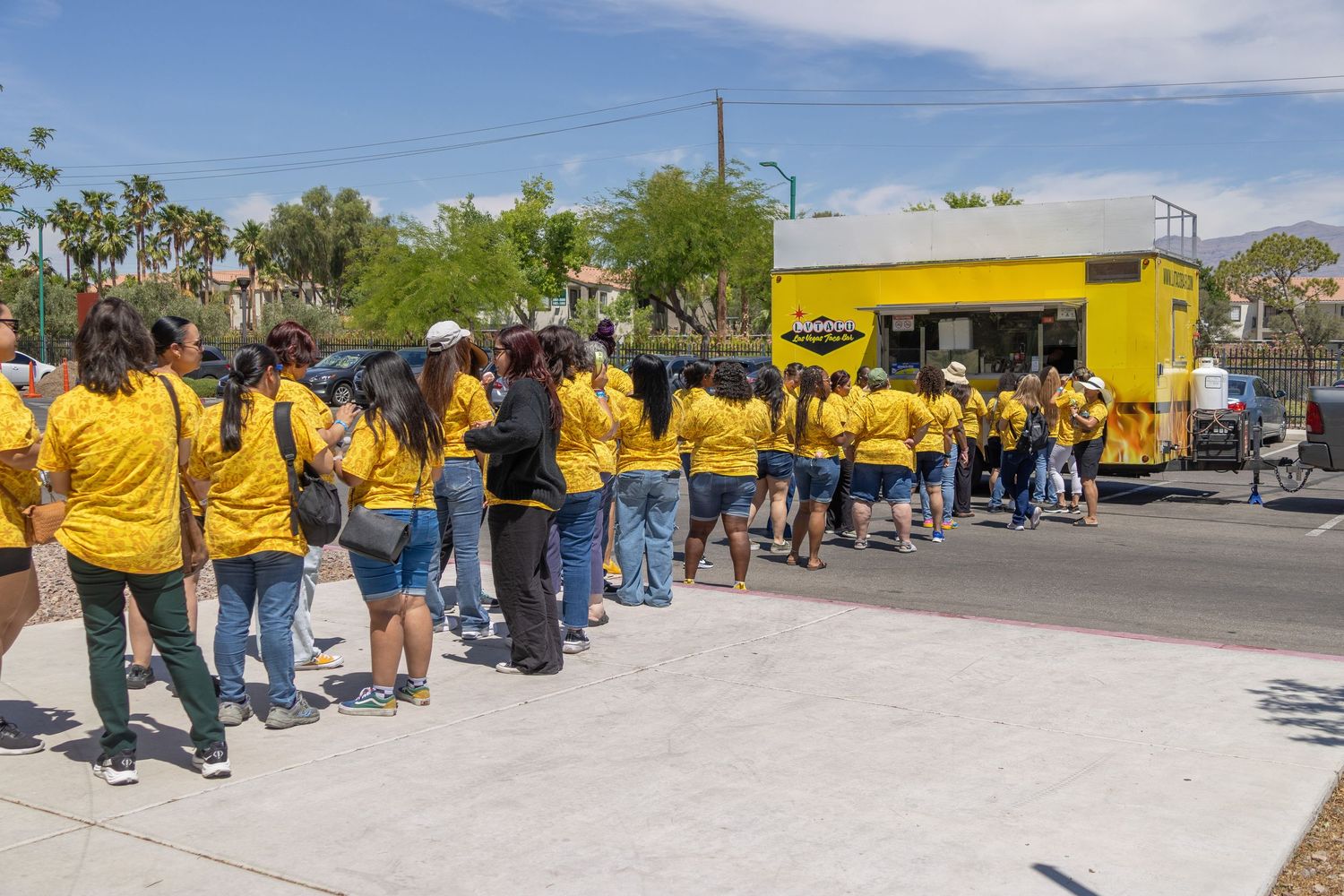 Large group of volunteers in yellow shirts wait in line outside a building on a sunny day.