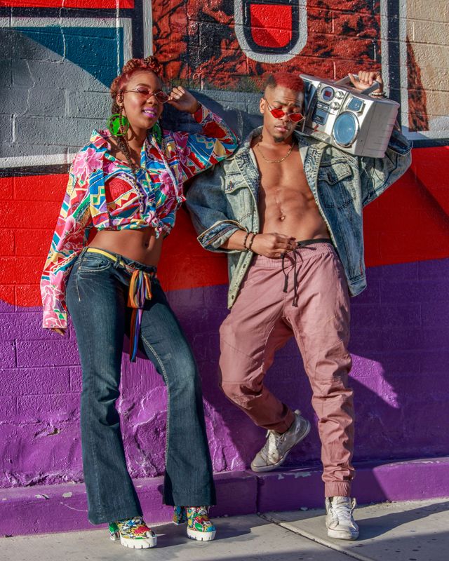 Two people lean against a colorful wall with purple and red sections while wearing casual urban streetwear.