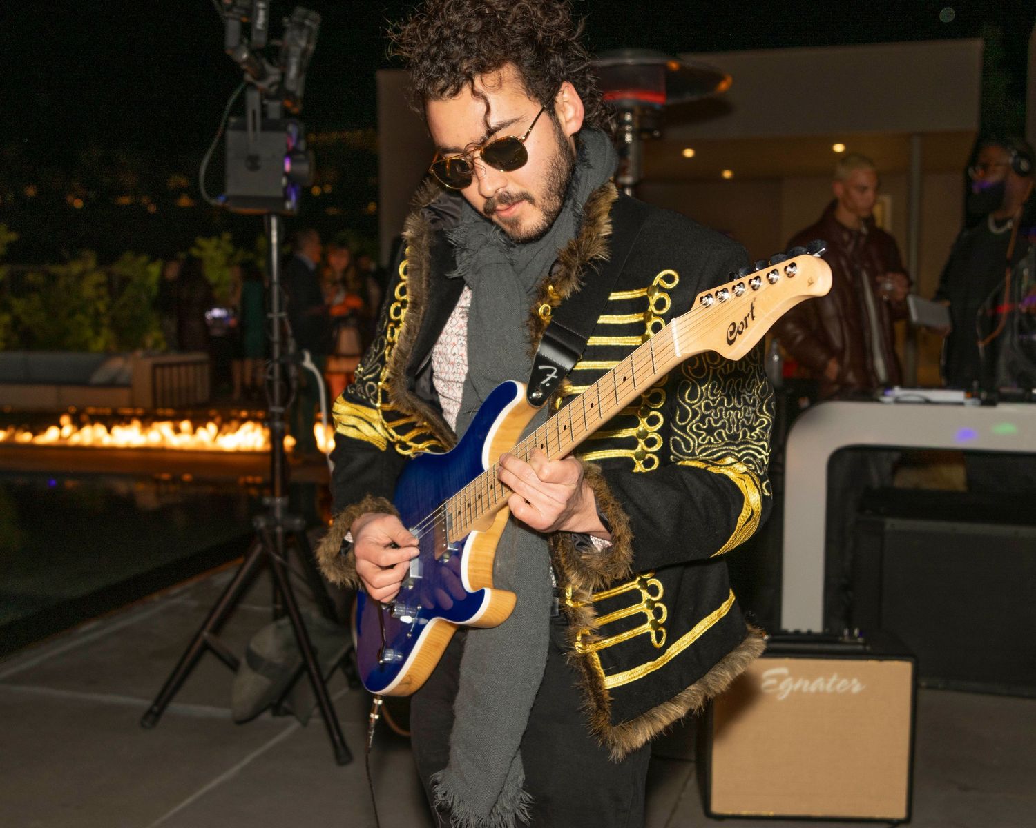 Musician playing guitar at outdoor venue with decorative lighting and crowd.
