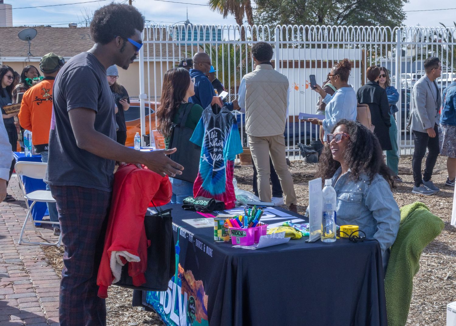 Outdoor community event with various vendor tables and booths set up along a path where people are walking and browsing.