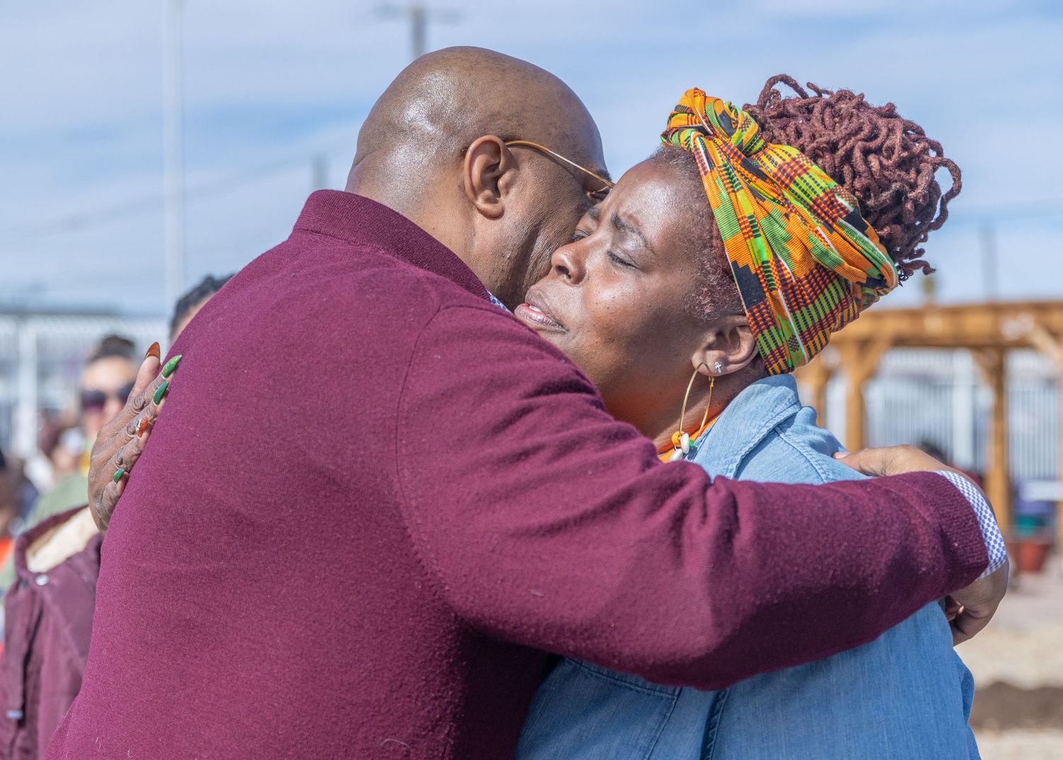 Two people sharing a tender embrace while dressed in burgundy and denim on a sunny day outside.