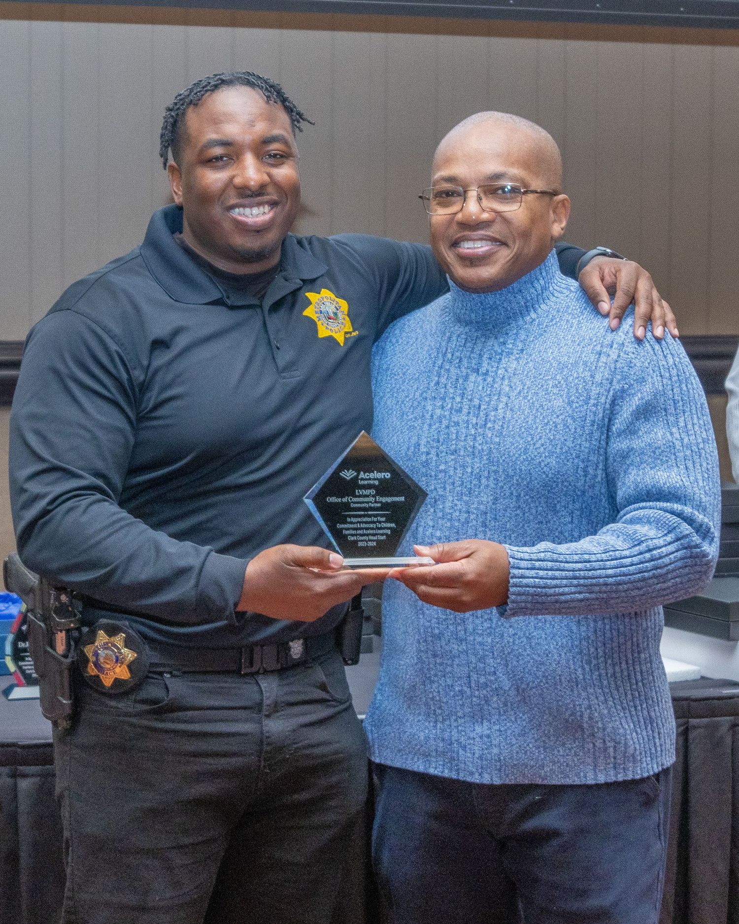 A person in a blue sweater receives an award from someone in law enforcement uniform at an indoor event.