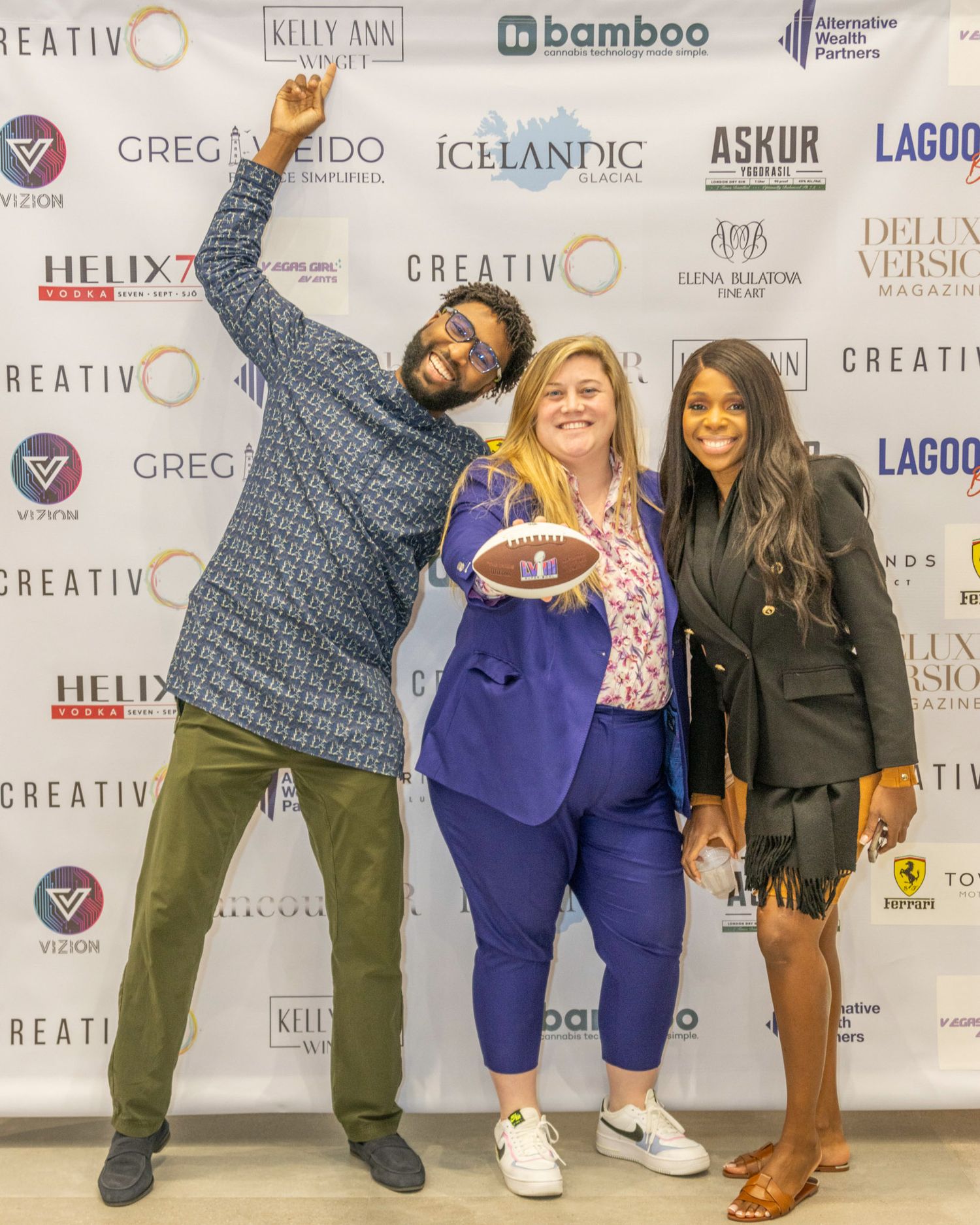Three people pose energetically in front of a sponsor wall with various company logos during an event.