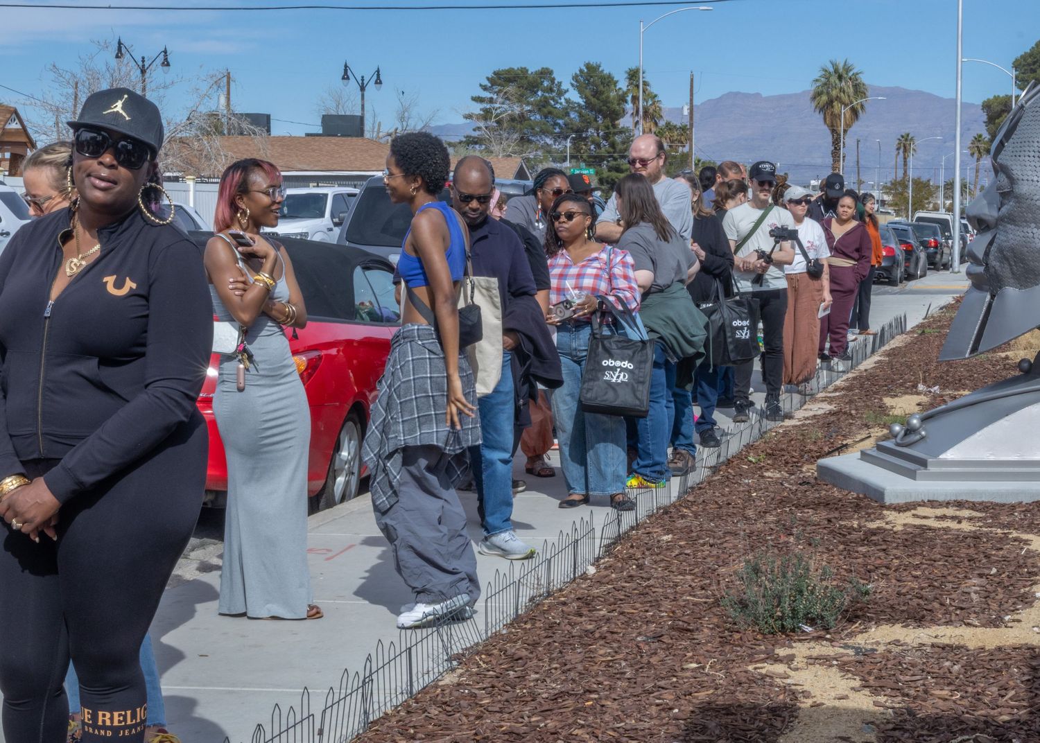 A diverse crowd gathers along a landscaped walkway with palm trees and mulched garden beds during a daytime event.