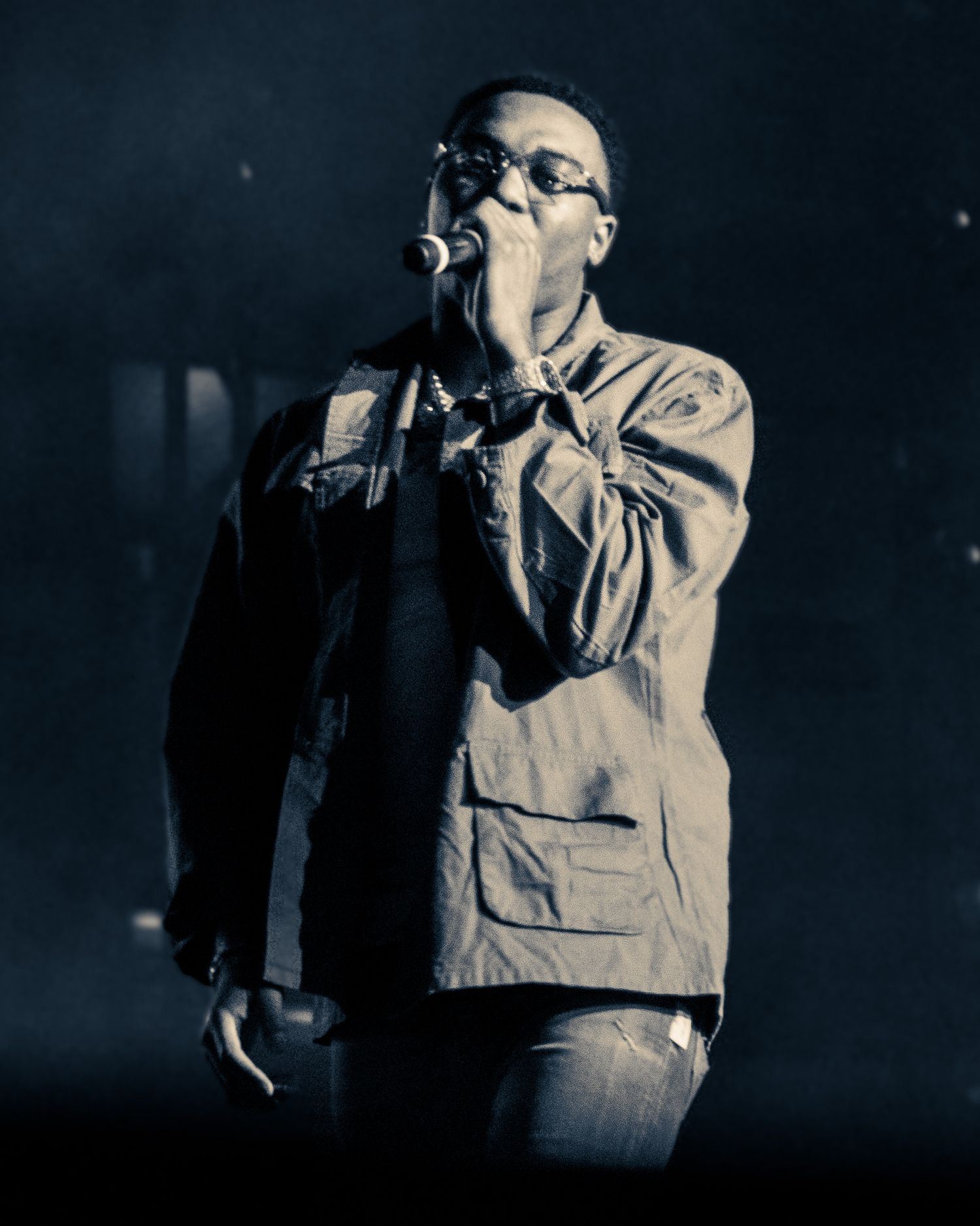 A performer in a beige jacket sings passionately into a microphone on a dark stage.