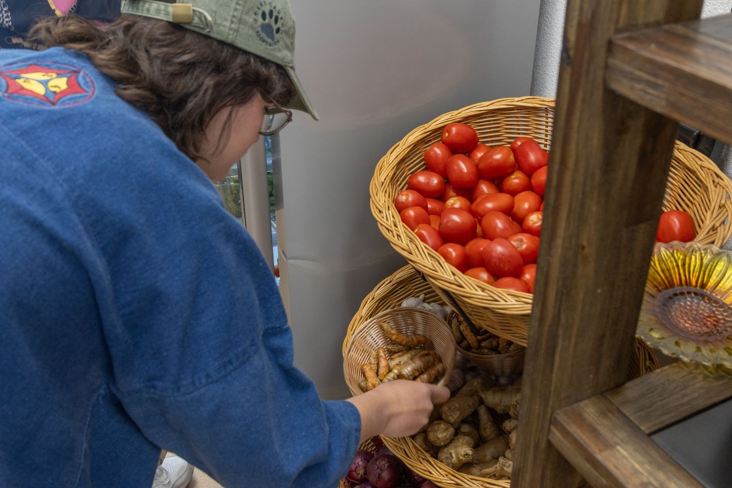 A person in a blue denim jacket selects produce from wooden shelves displaying fresh tomatoes and potatoes.