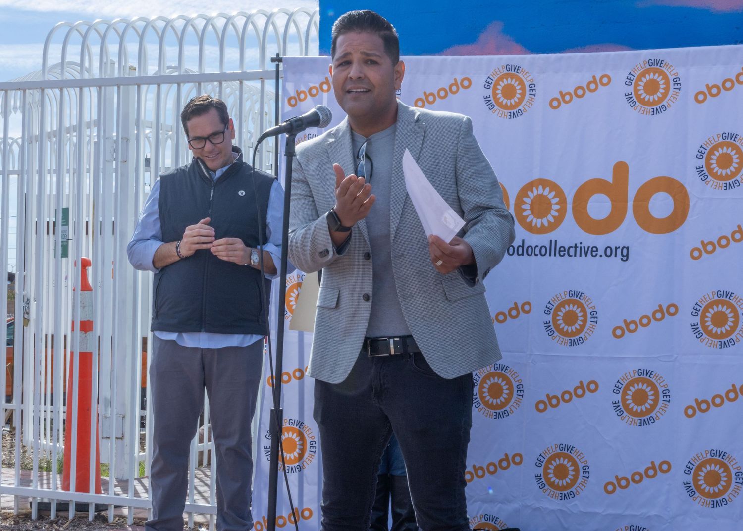 A series of speakers stand at podiums against a backdrop with repeating orange circular logo designs.