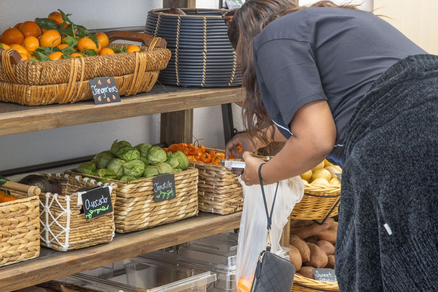Someone arranges fresh produce in wicker baskets on wooden shelves at a local market display.