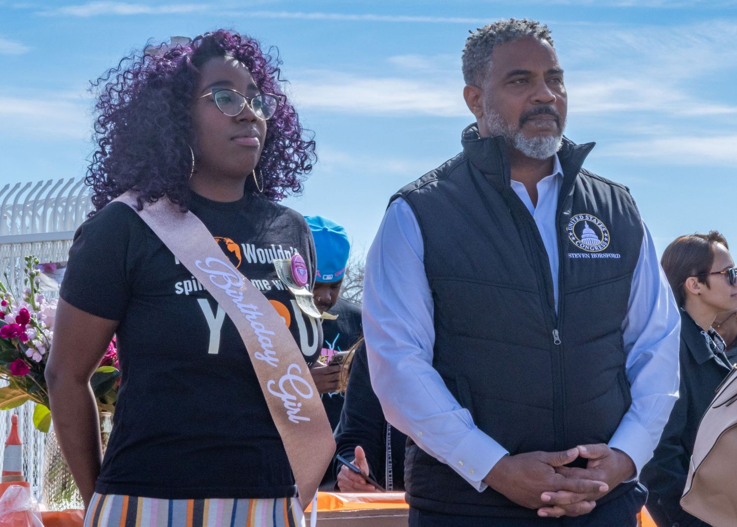 A group gathers outdoors at a community event wearing name tags and colorful outfits on a sunny day.