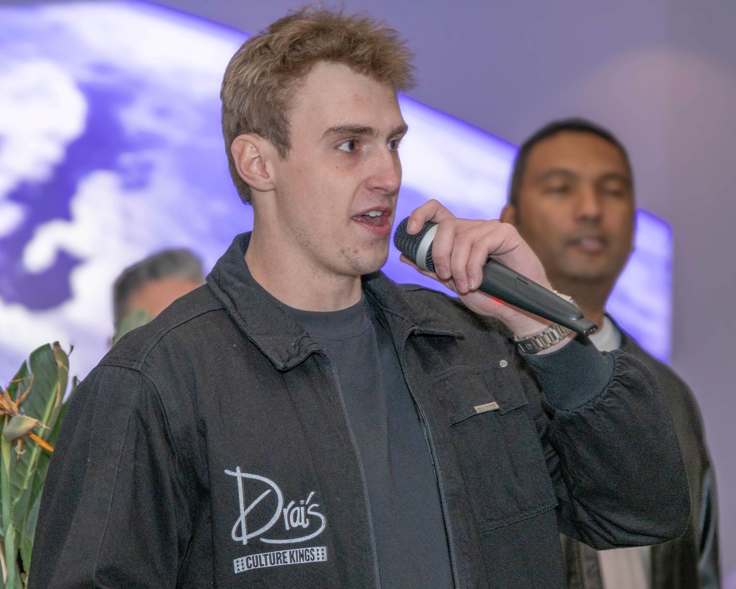 Performers on stage with microphones during an indoor event with purple lighting.