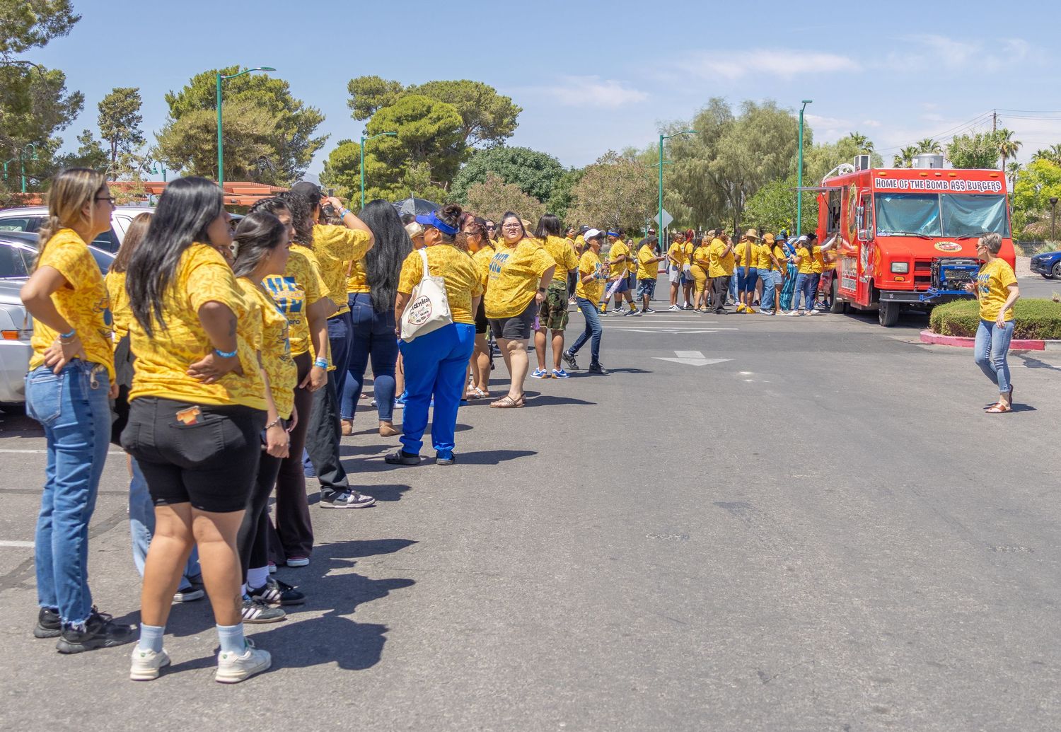 Panoramic view of crowds gathered in a parking lot during an outdoor community event on a sunny day.