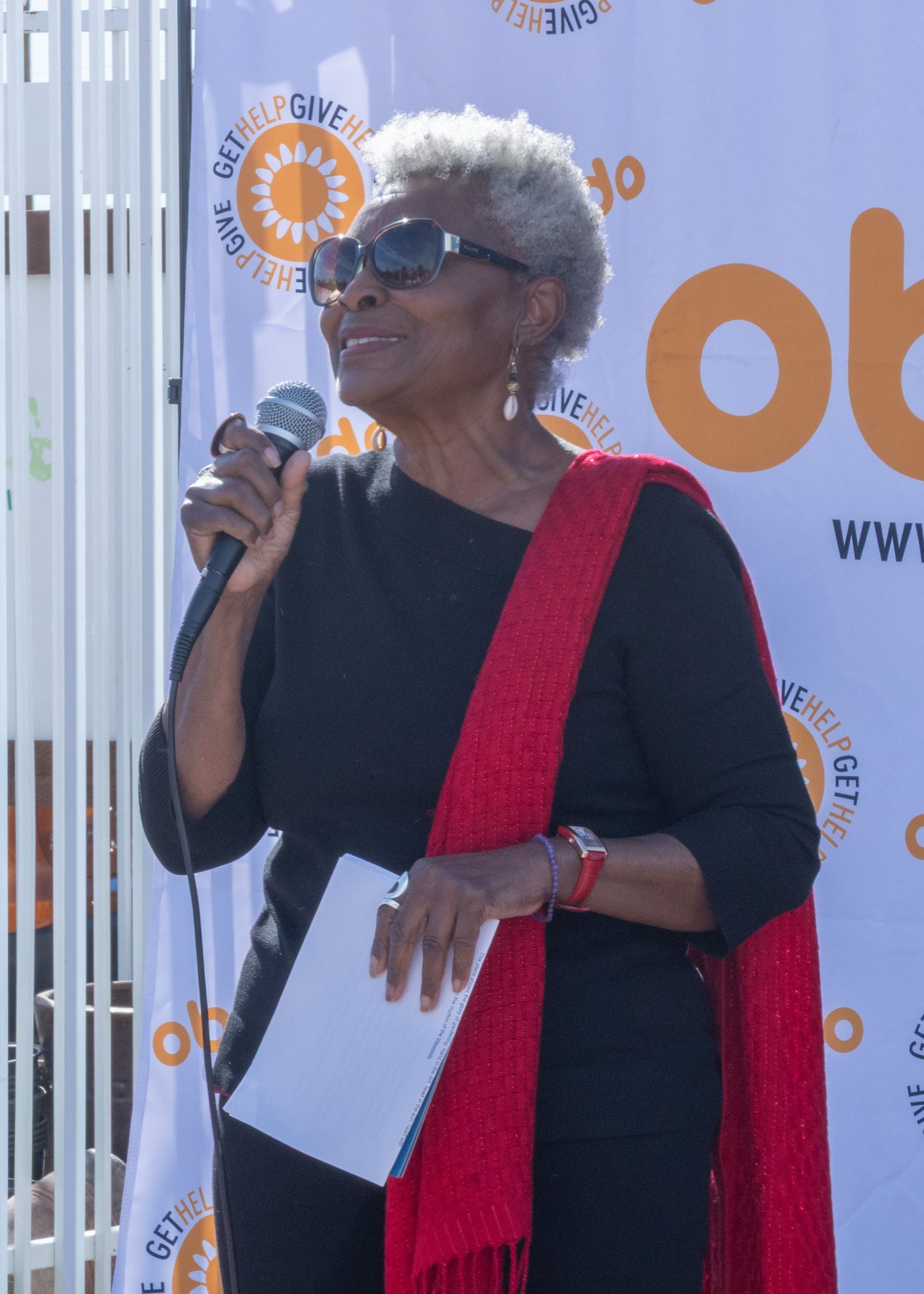 Someone speaks into a microphone at an event wearing a black dress with a red scarf against an orange and white backdrop.