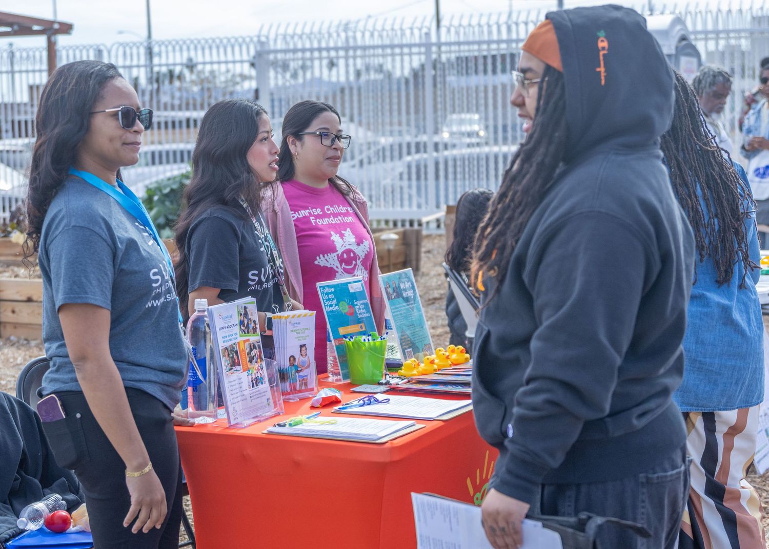 People gather around tables at an outdoor community event with drinks and food on display.