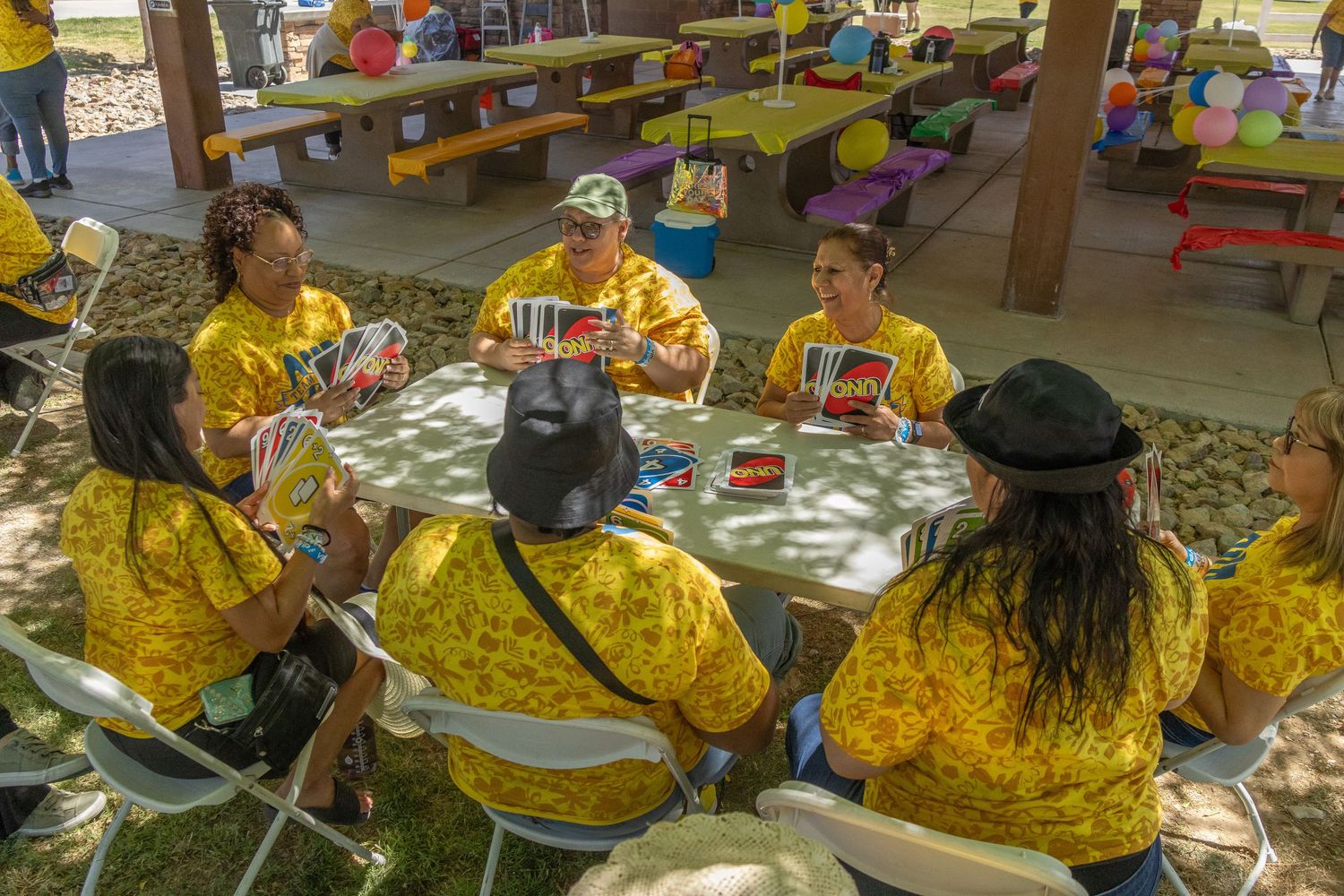 Community gathering with people seated at picnic tables under a shaded pavilion.