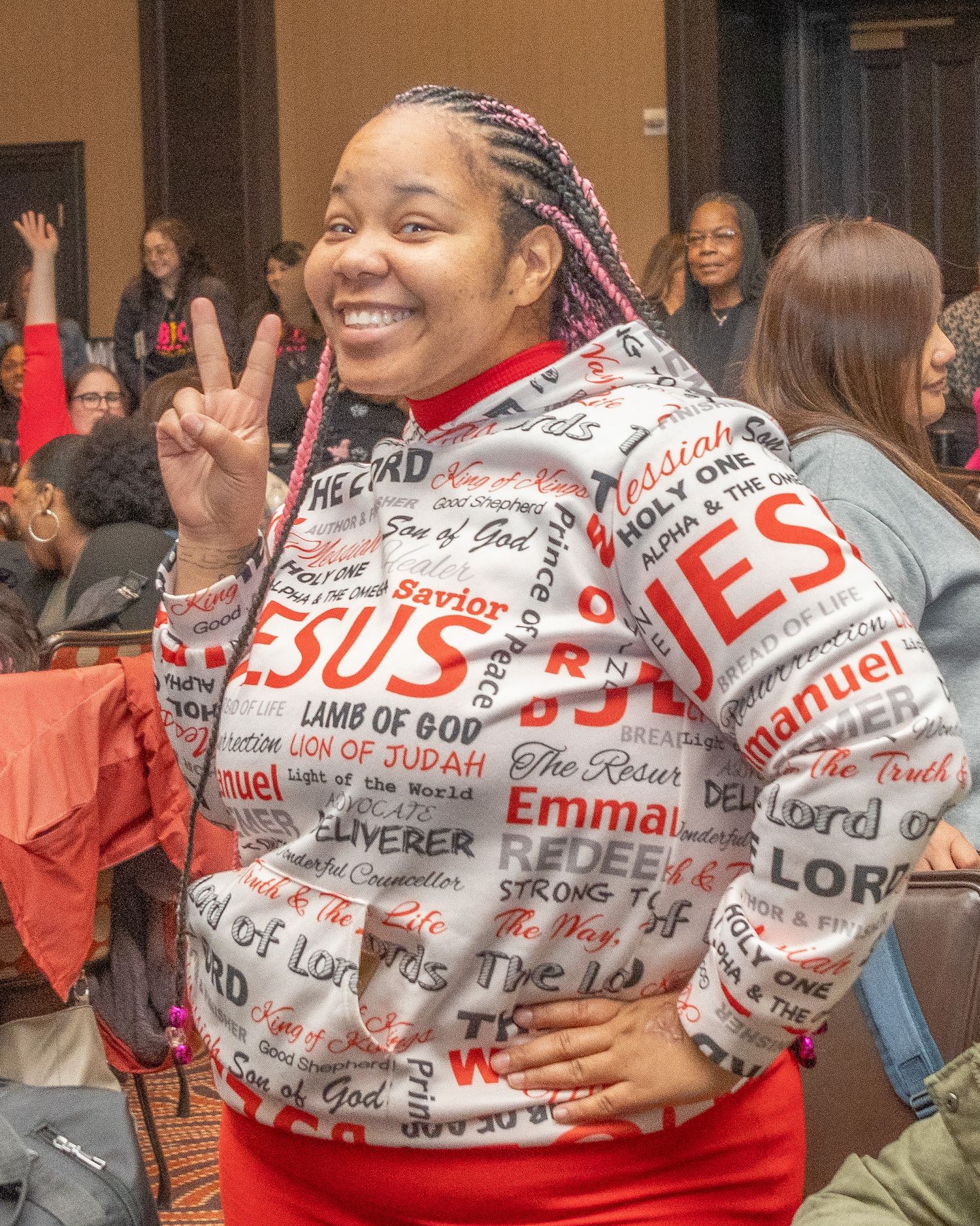 Enthusiastic attendee wearing a religious-themed sweater with text design makes peace sign at event.