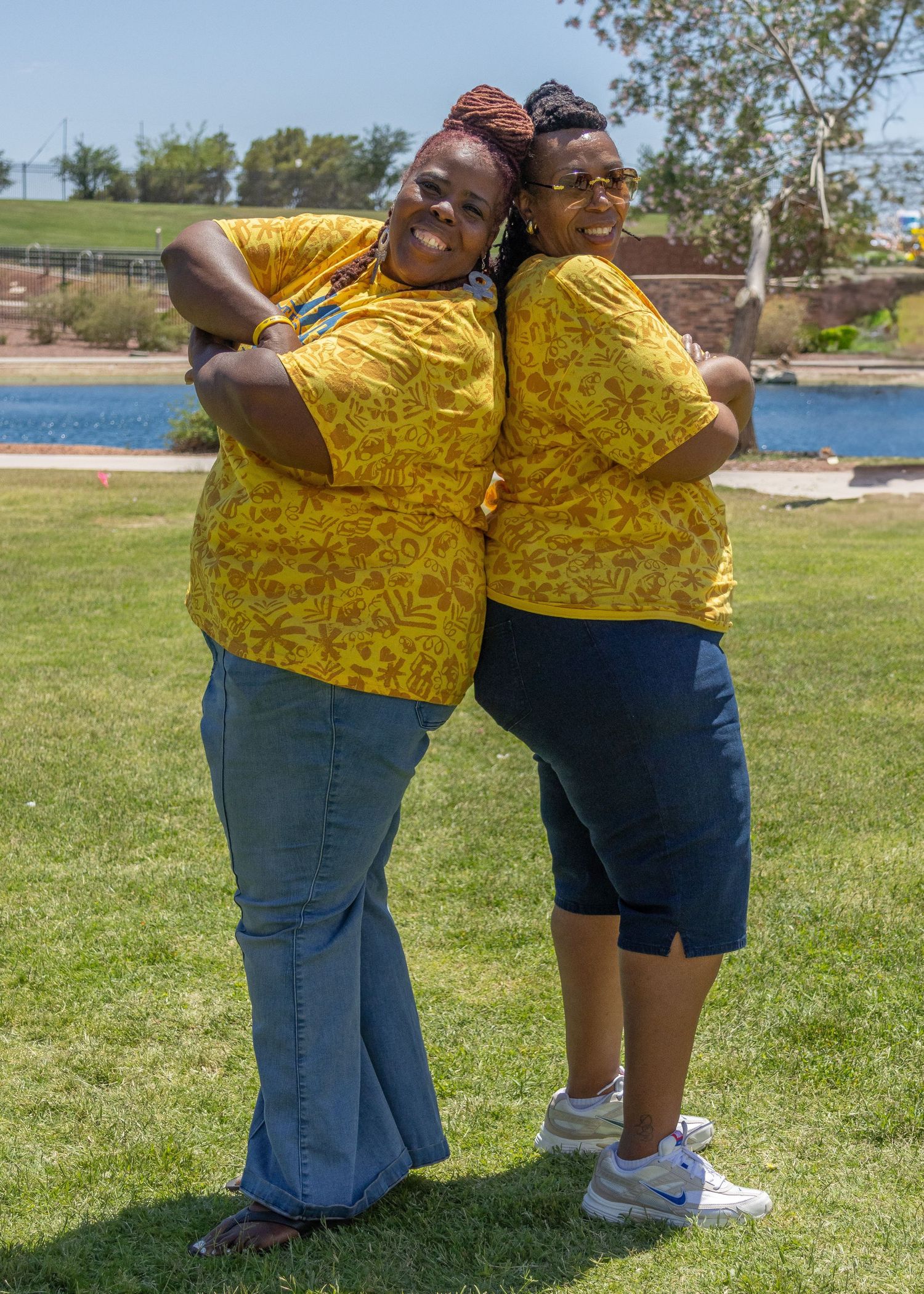 Two individuals wearing matching yellow floral shirts and blue bottoms stand back-to-back outdoors,  Tameka Henry. Acelero Learning.