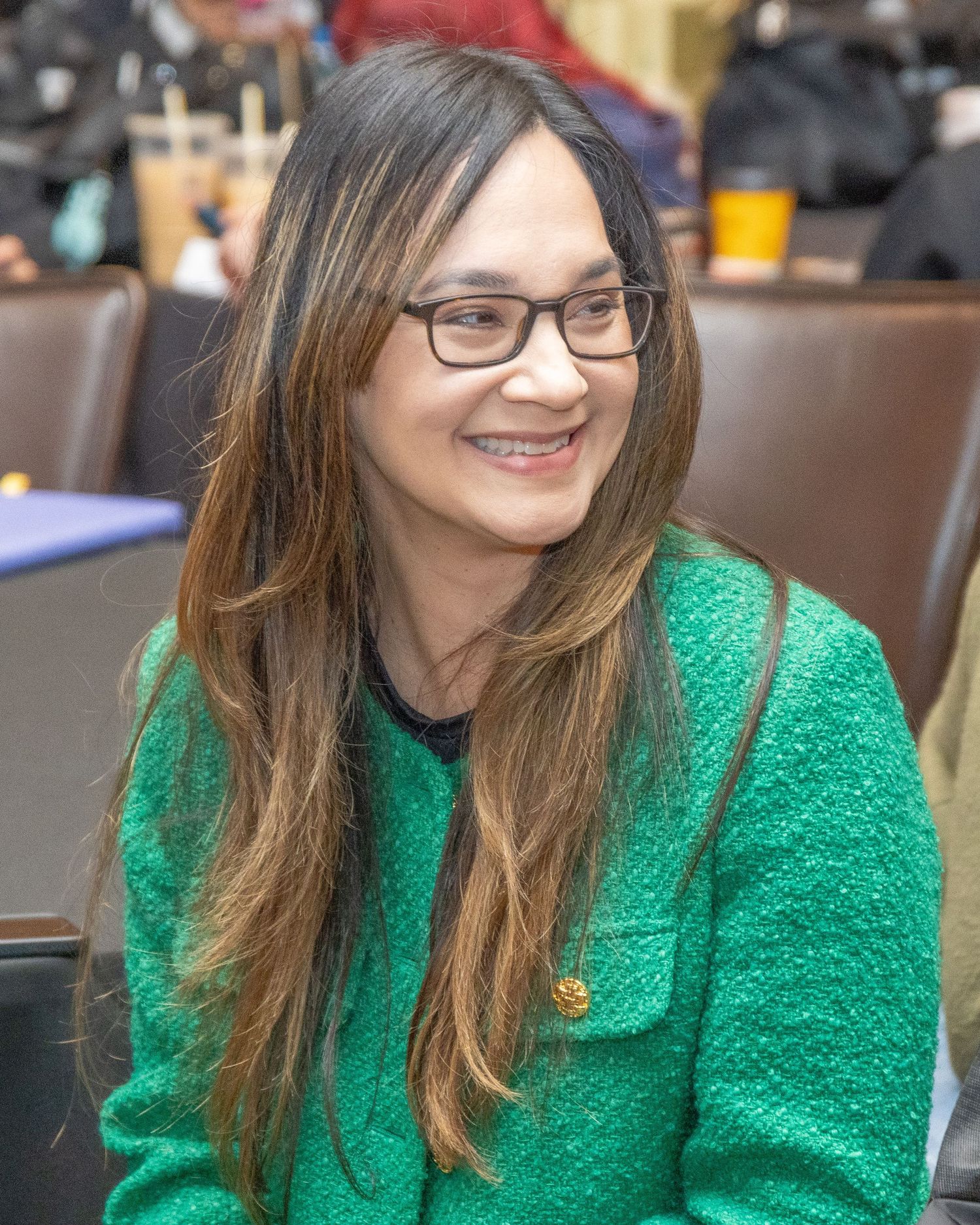 Person in green sweater smiles warmly while seated at indoor event.