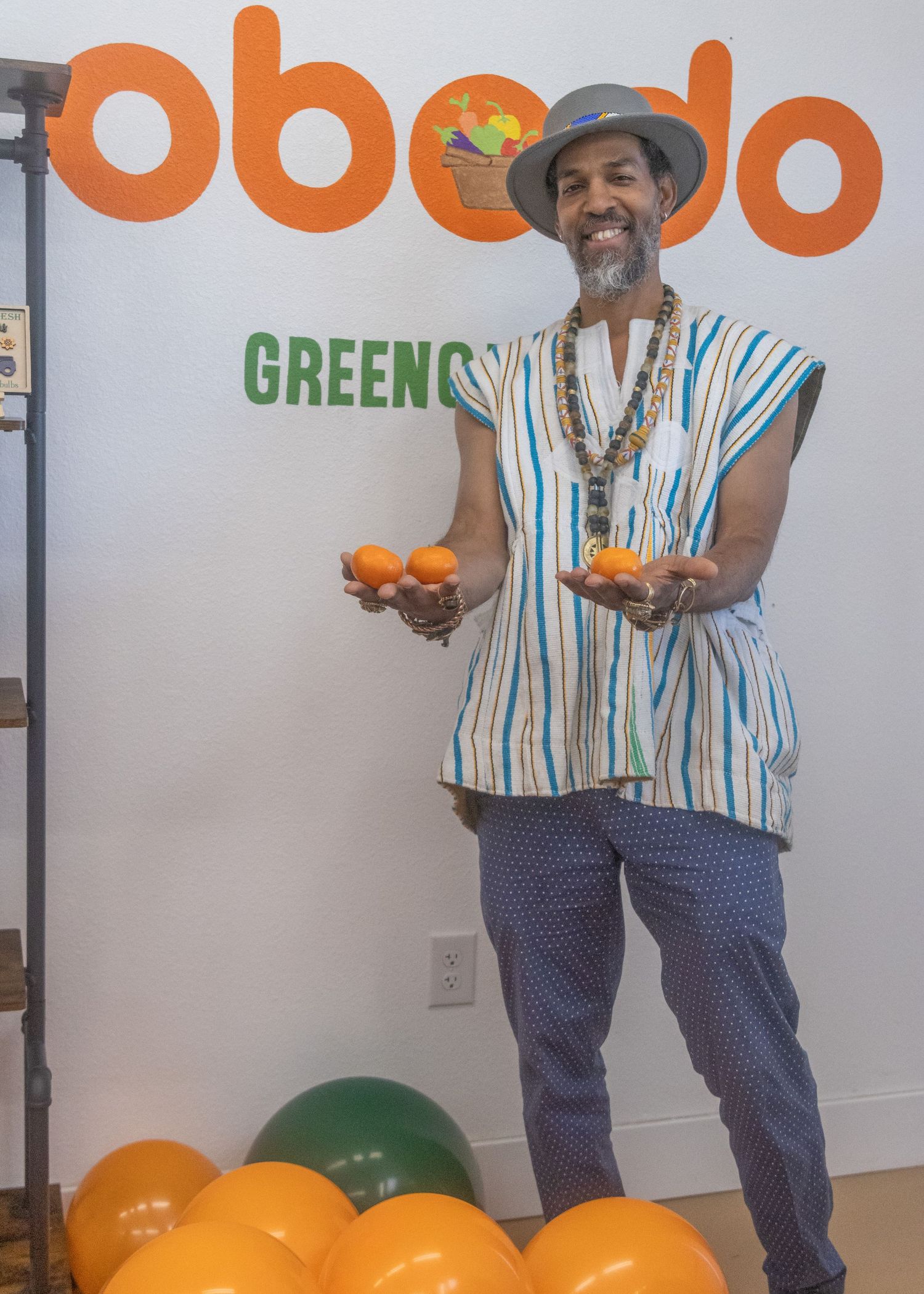 Someone in a striped shirt and navy pants poses with oranges and green and orange balloons in an office setting.