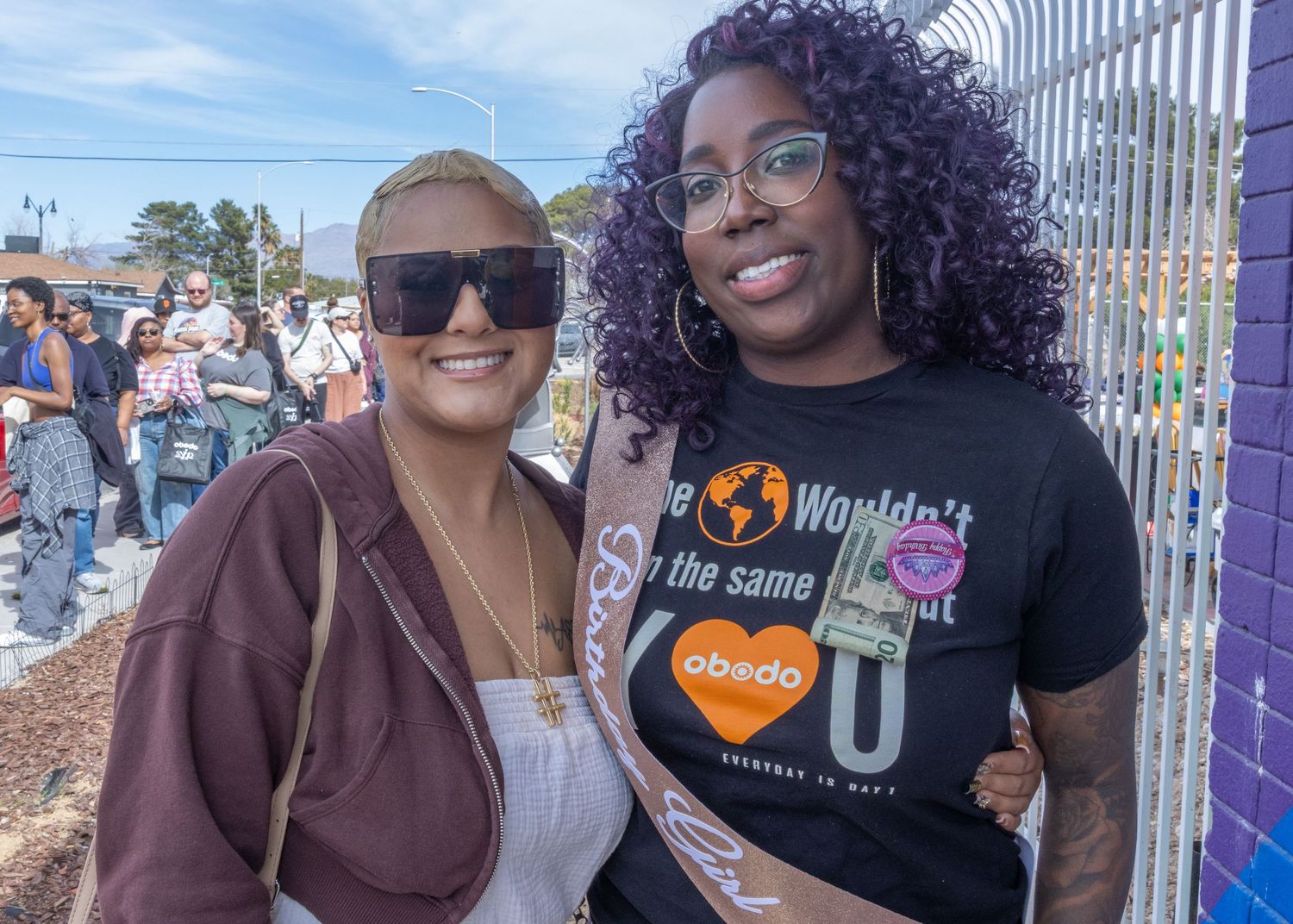 Two people smile together outdoors wearing casual outfits and sunglasses during a sunny day.