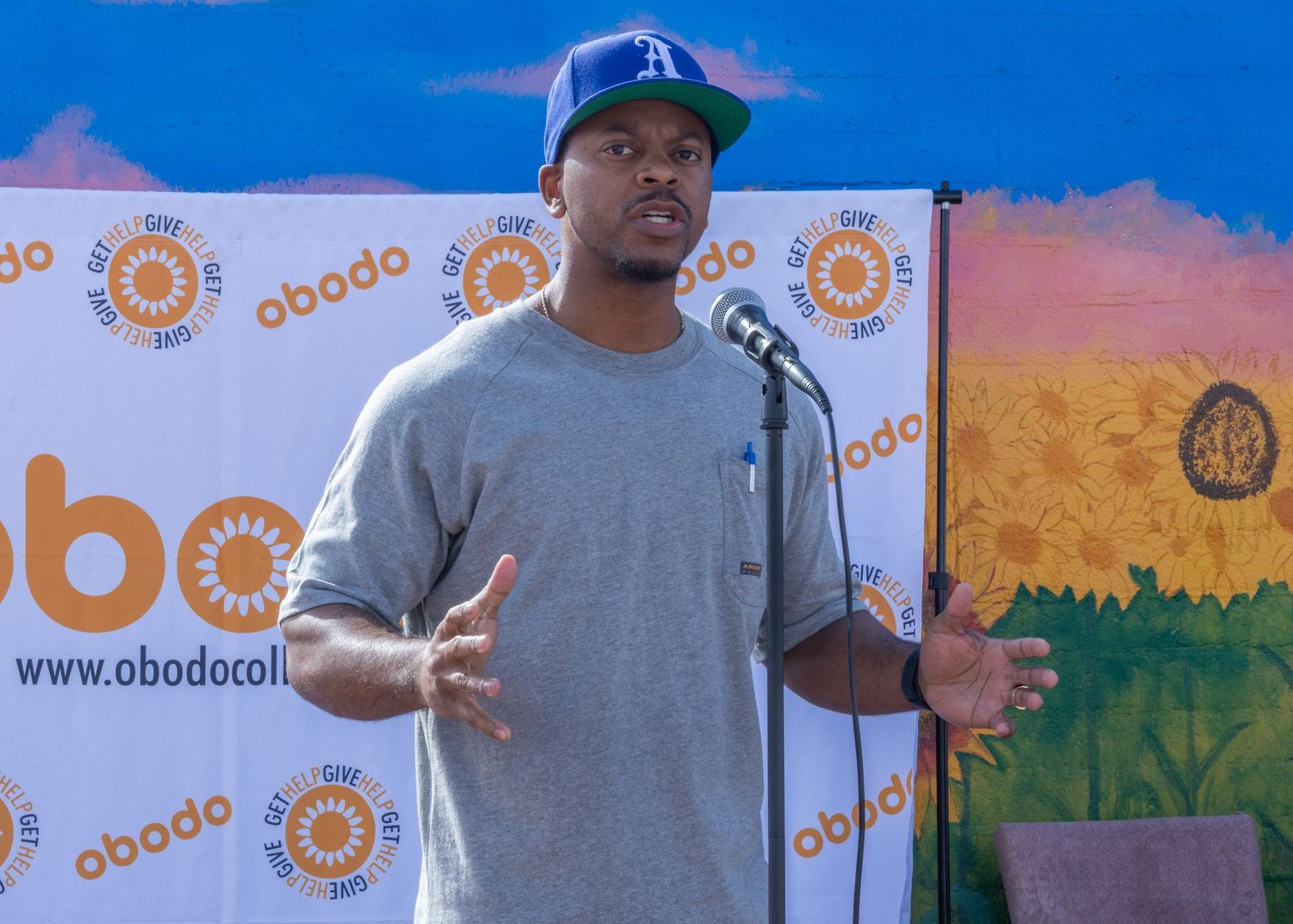 A person in a baseball cap stands in front of a backdrop with orange circular logos during an event presentation.