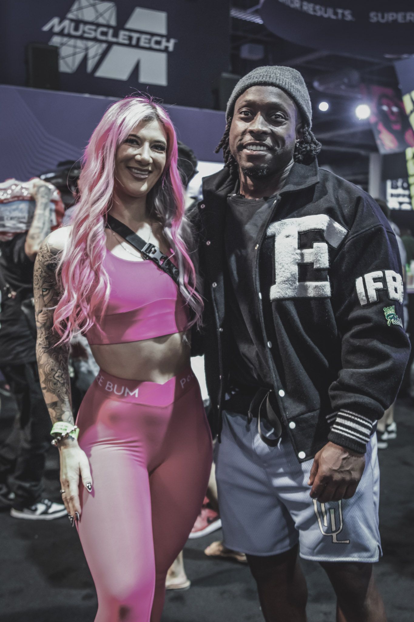 Two people posing together at a fitness expo or event, with one wearing pink athletic wear and another in dark clothing.