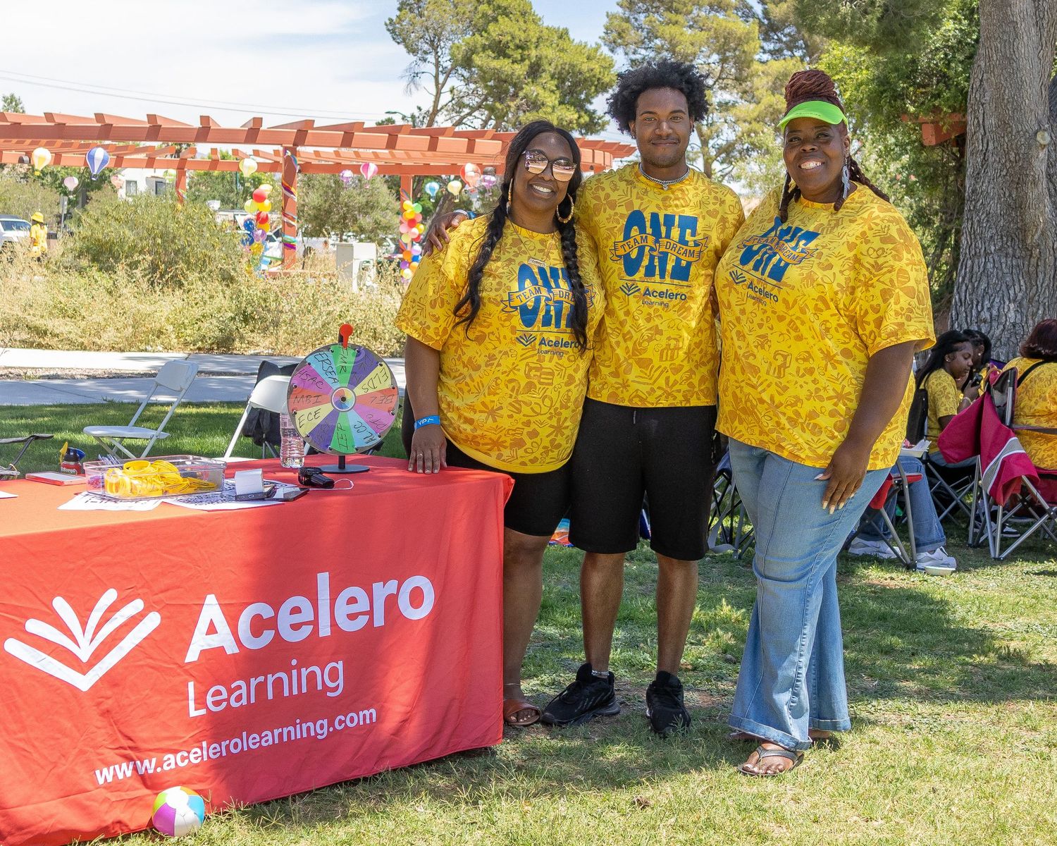 A group of people wearing yellow shirts gather outdoors near event banners and signage. Acelero Learning