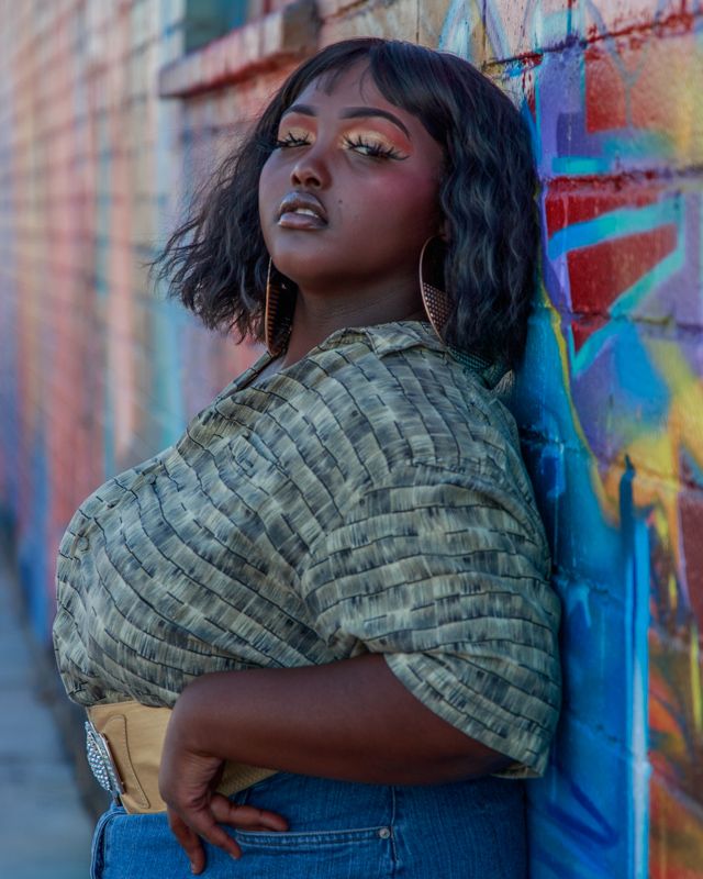 Portrait against colorful graffiti wall showing dramatic eye makeup and a textured green top paired with denim.