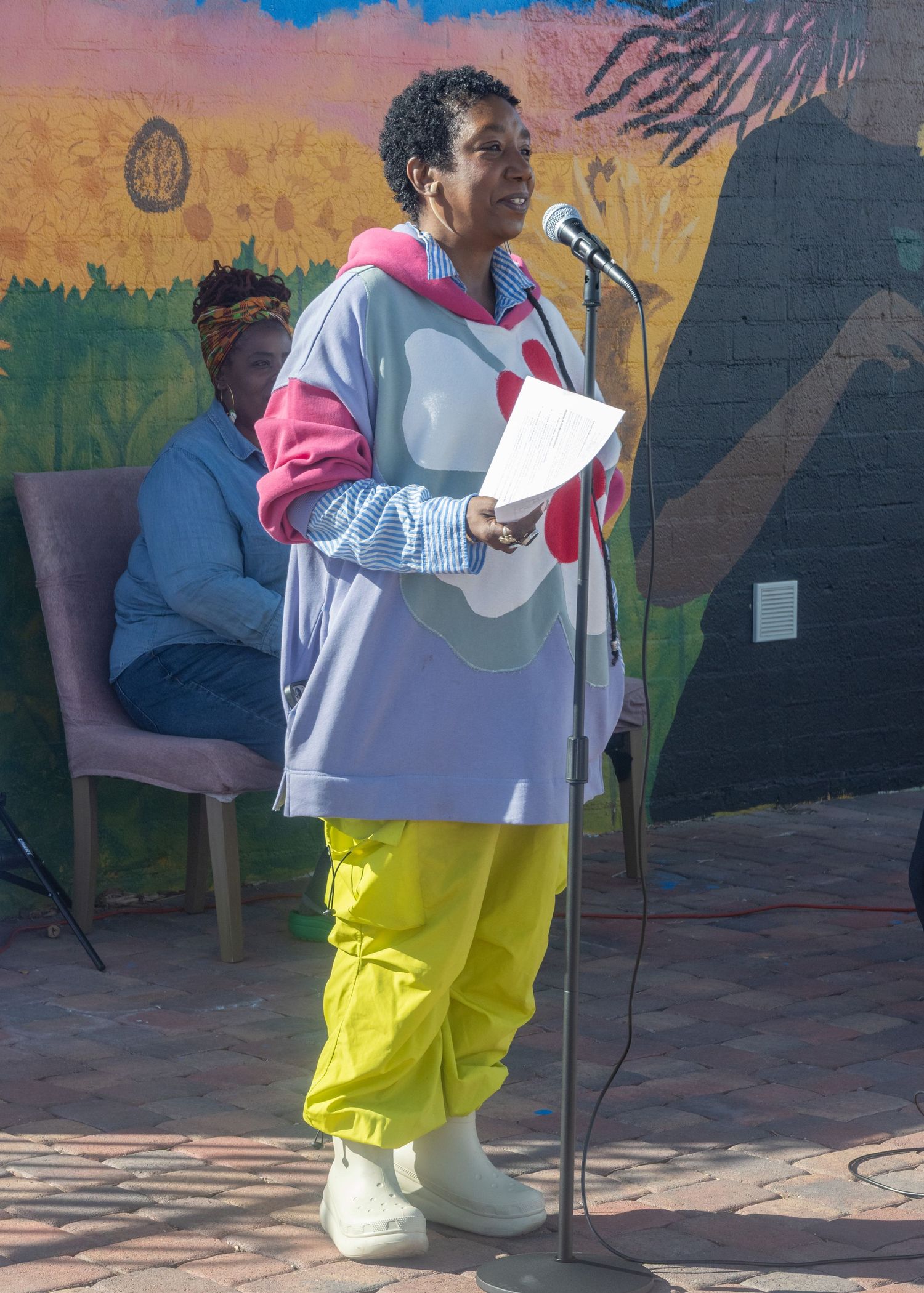 Person sings into microphone wearing yellow pants and striped top against colorful mural backdrop.