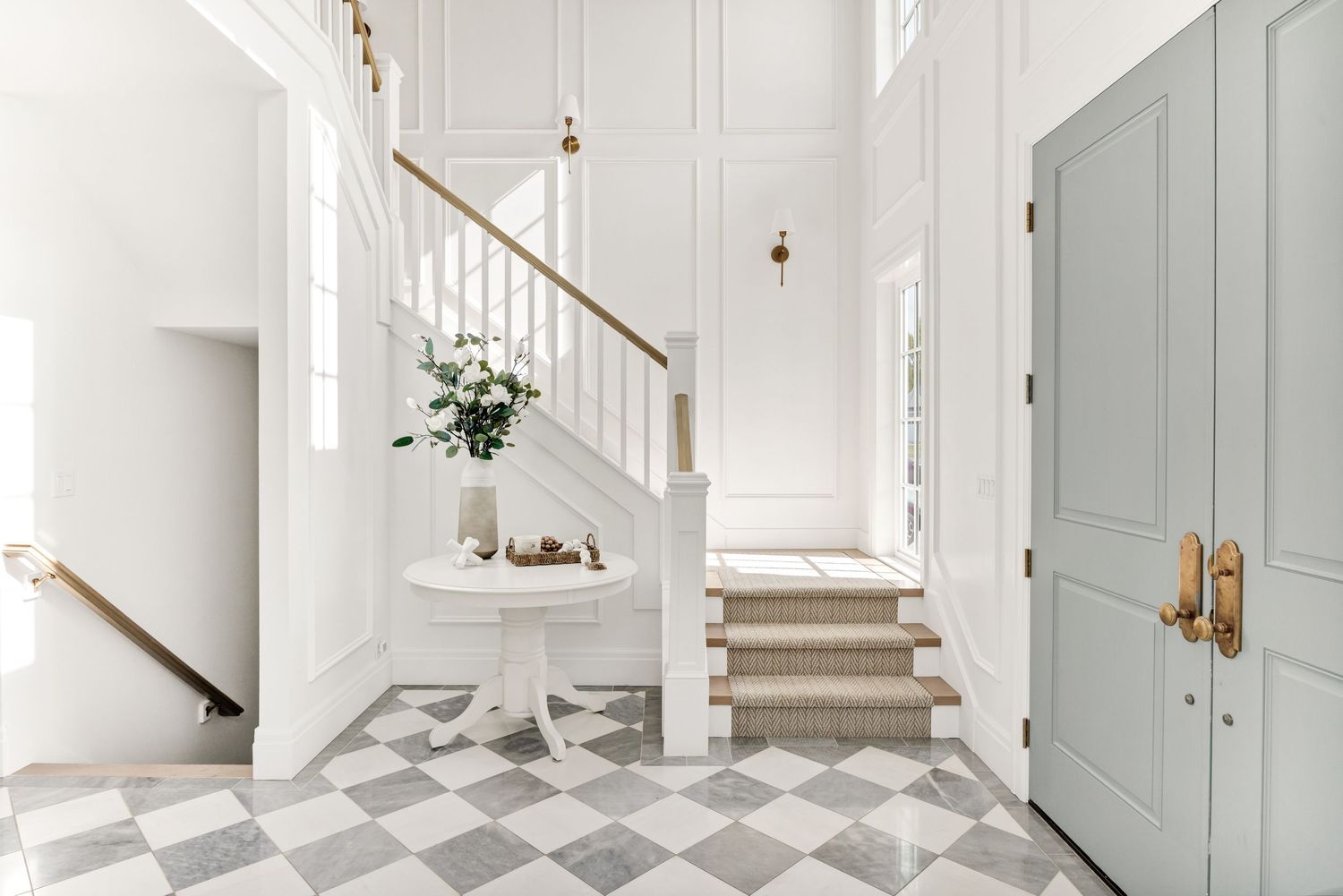 Bright white entryway with checkered marble floor, elegant staircase, round accent table, and a sage green front door with brass hardware.