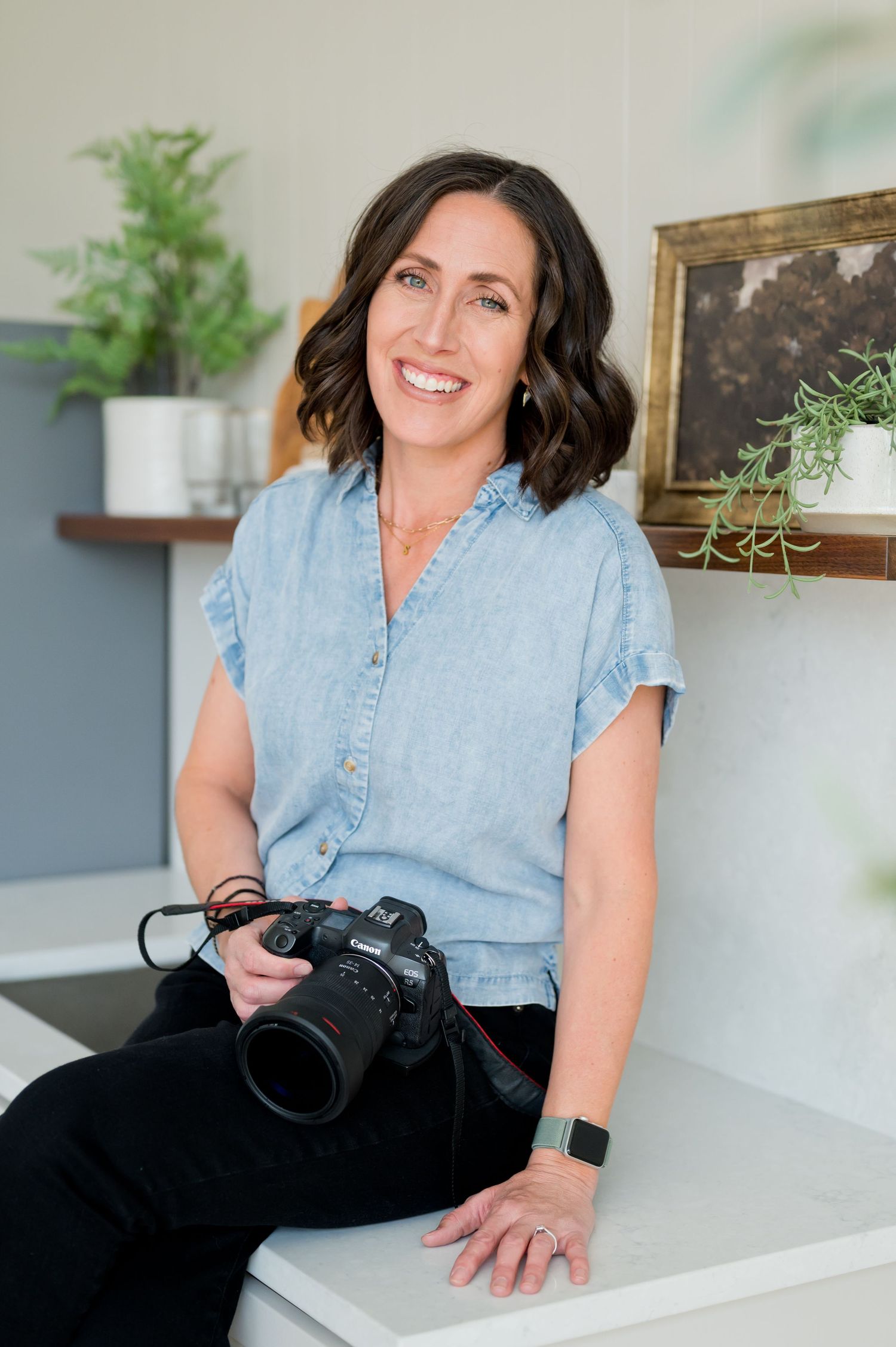 Photographer in denim shirt sits casually with professional camera in bright home office environment.