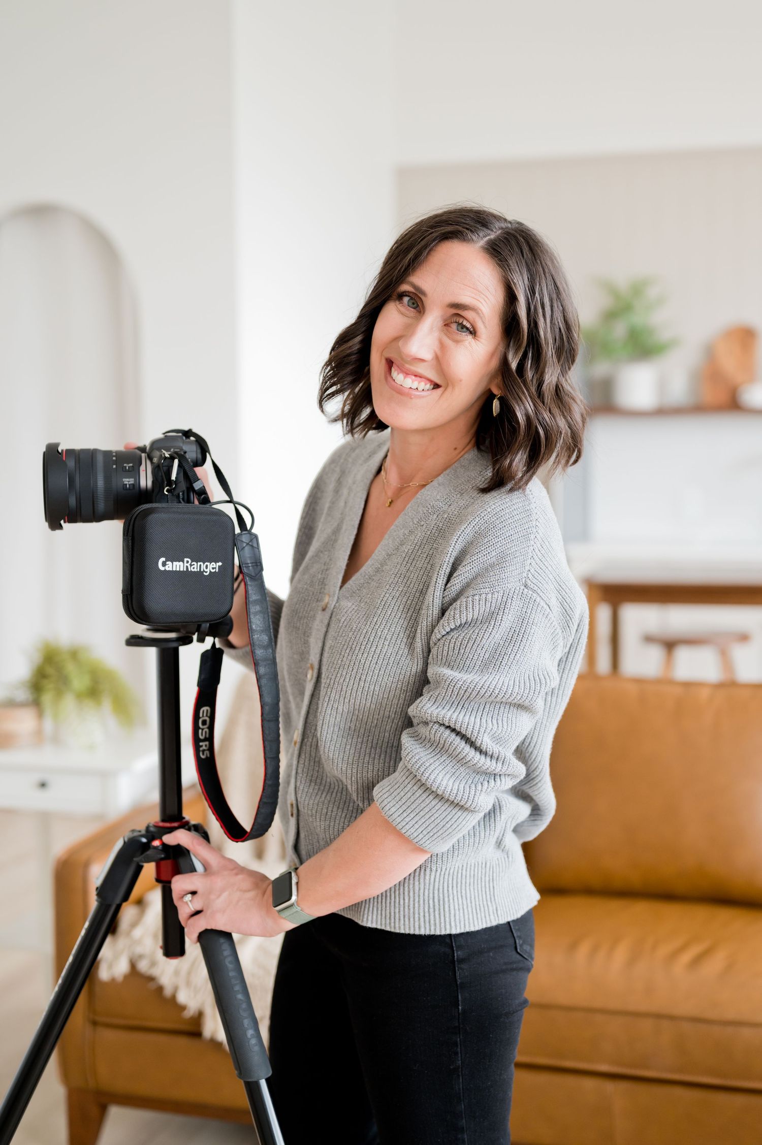 Photographer in grey sweater stands with professional camera mounted on tripod in modern living room.