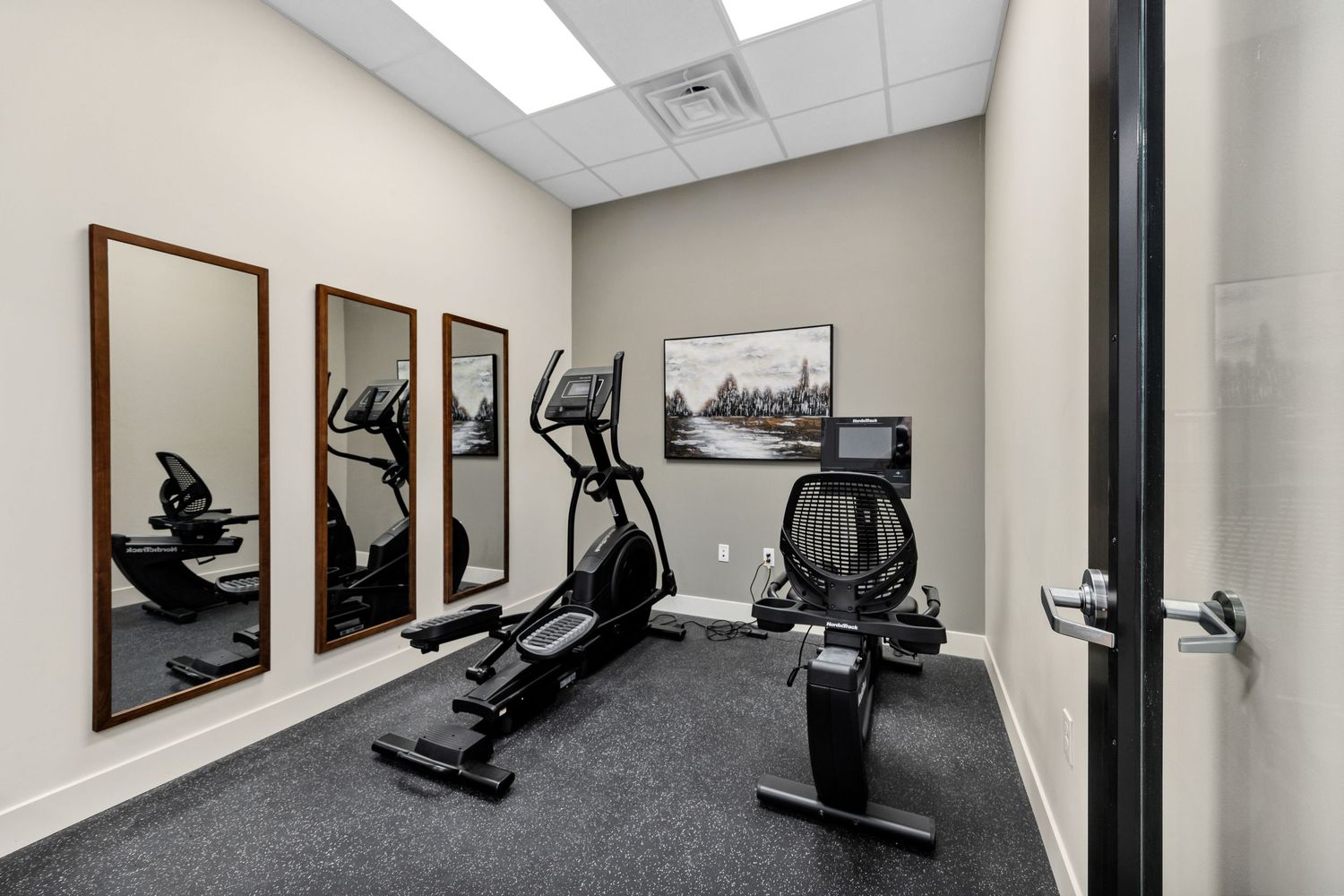 Exercise room with cardio equipment and framed artwork on beige walls.