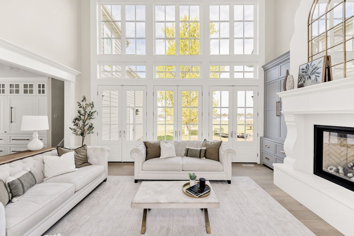 Bright white living room with two-story windows, white sofas, fireplace, and natural light flooding through French doors.