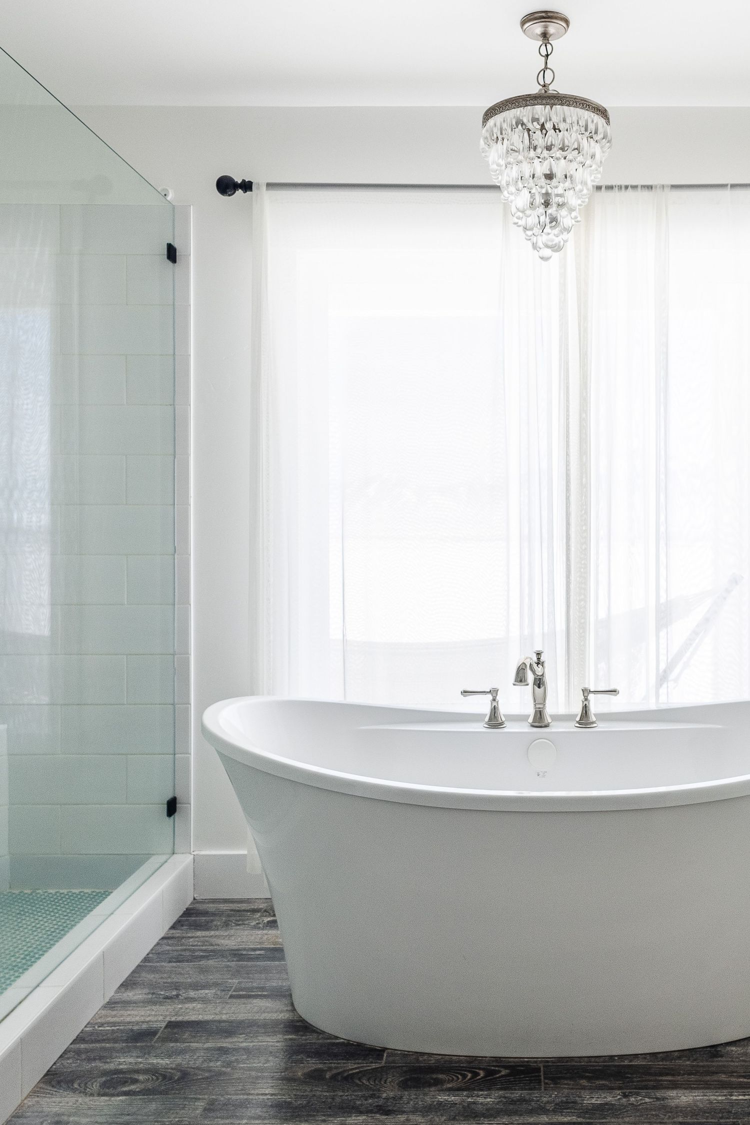 Modern luxury bathroom with freestanding white tub, crystal chandelier, and light blue glass wall panels.