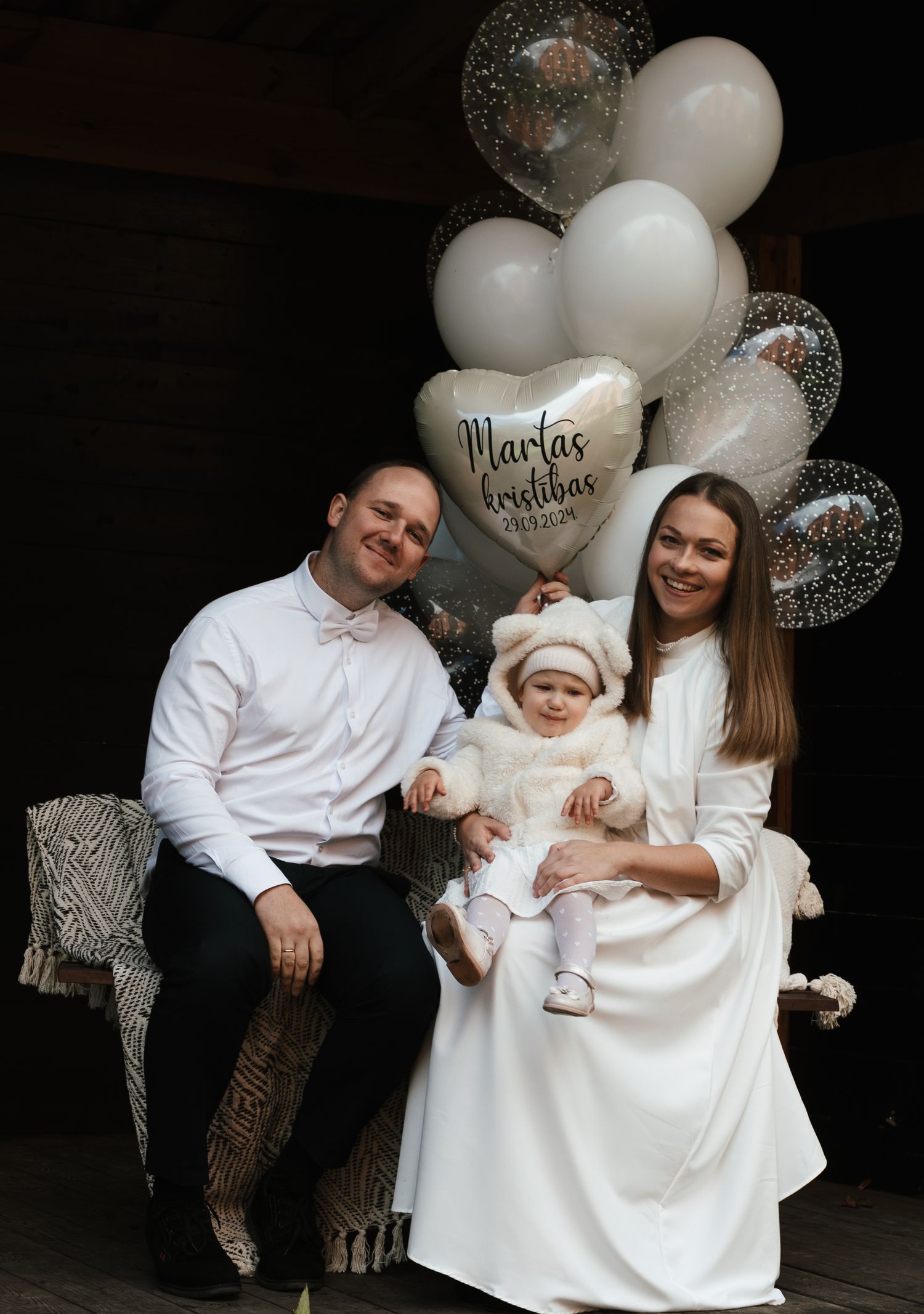 Vecāki sēž uz sola kopā ar meitu, aiz muguras viņiem daudz baltu balonu.