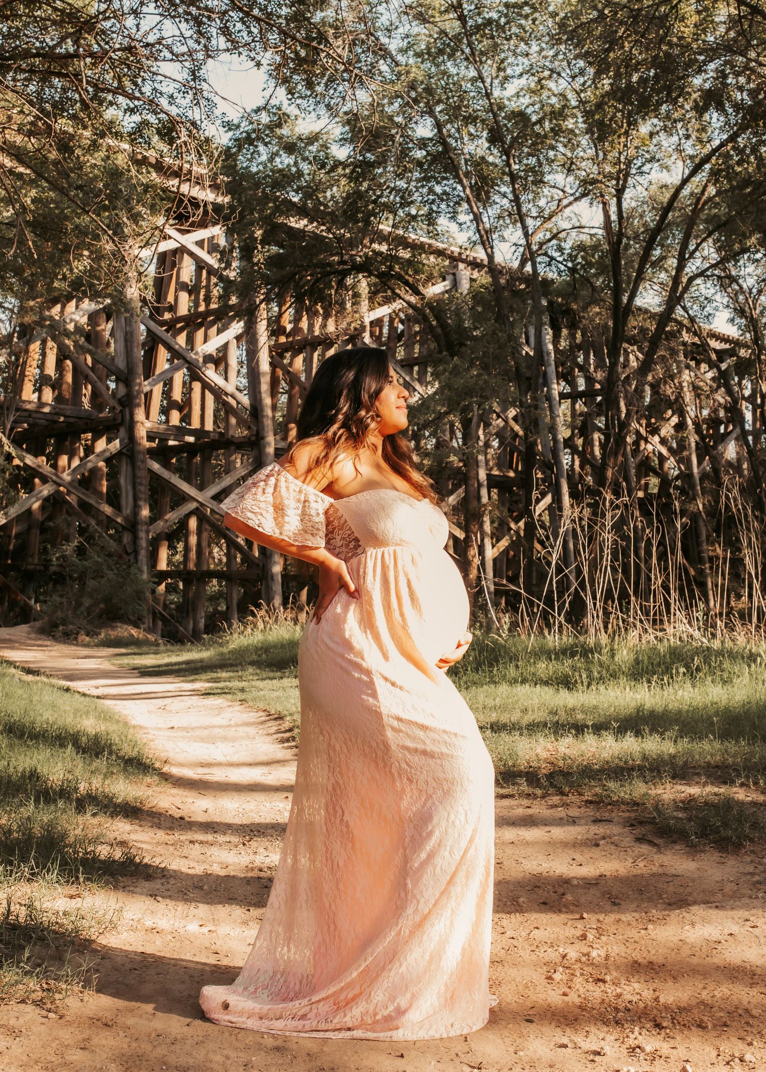 Pregnant woman in a flowing blush lace gown poses on a woodland path with a rustic wooden bridge backdrop.