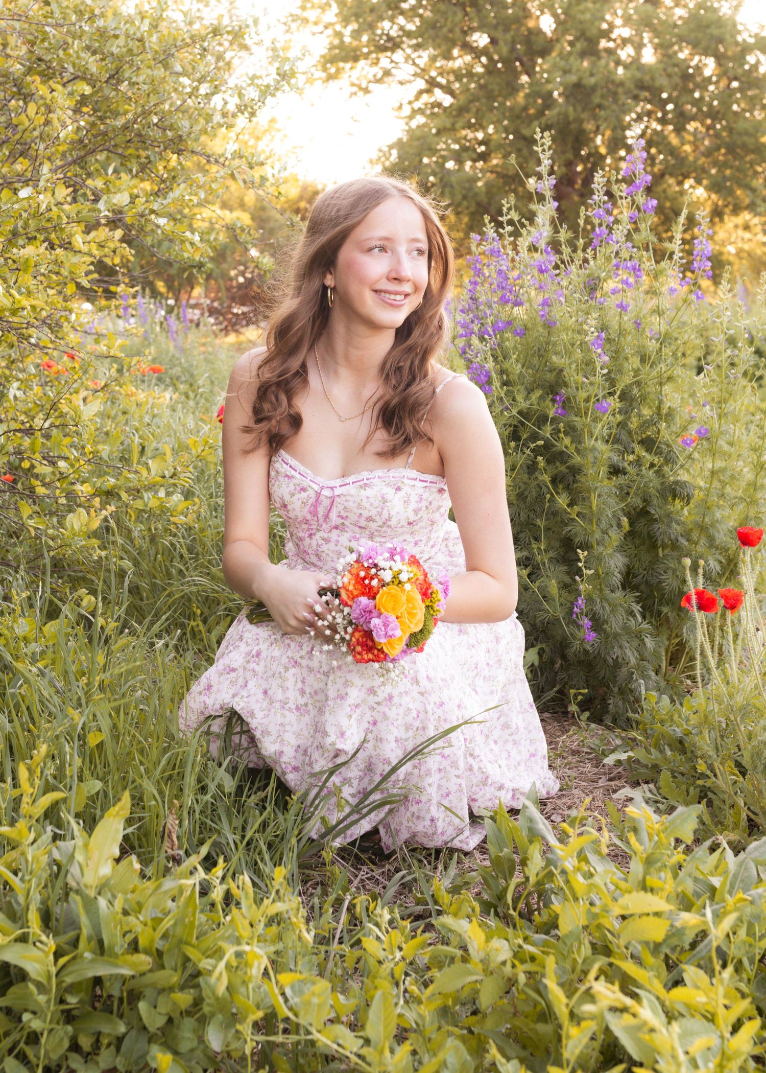 Young woman in floral dress holding colorful bouquet, kneeling in lush garden with wildflowers and purple blooms.
