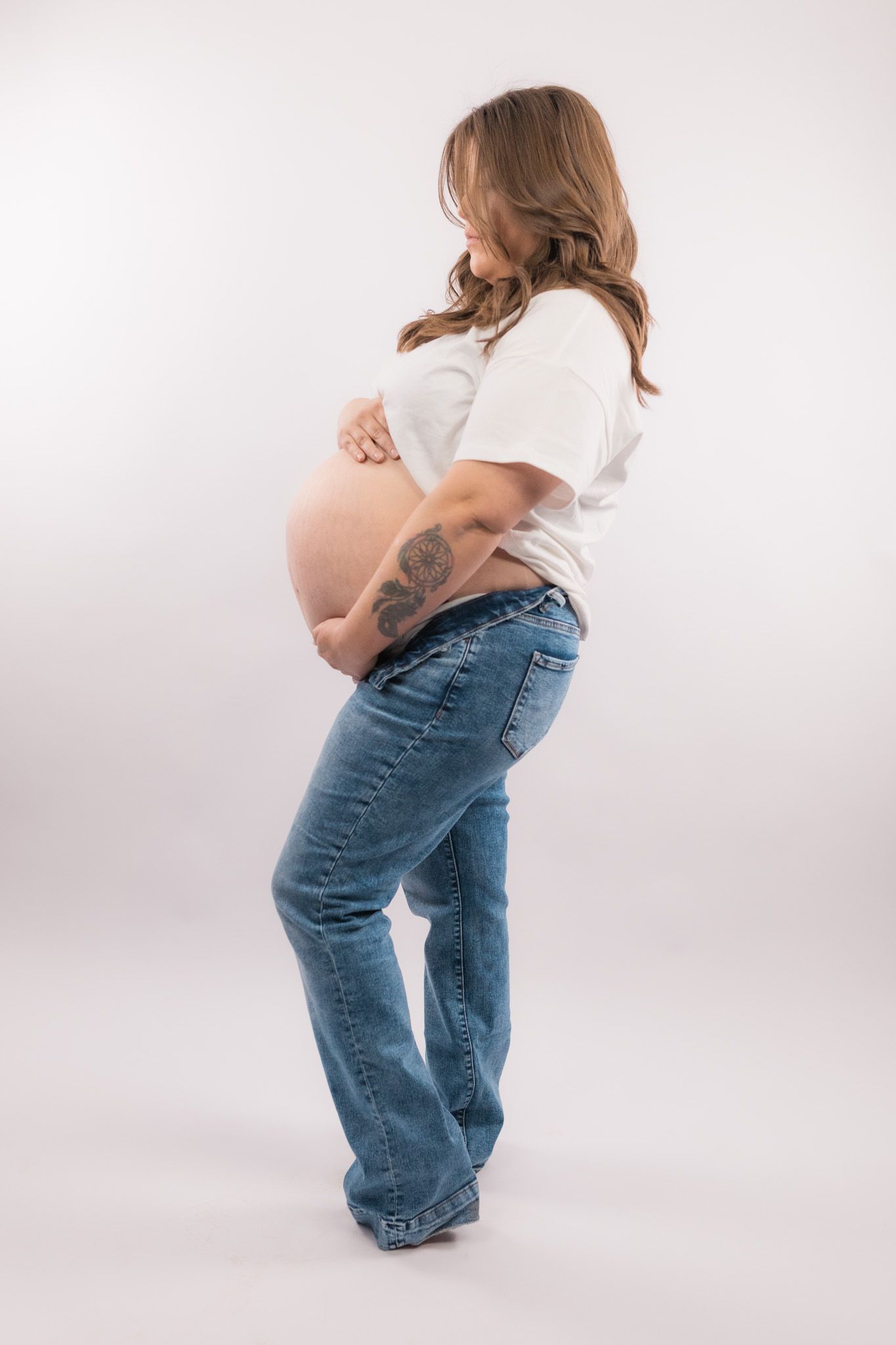 Artistic maternity photo showing a pregnant woman in white shirt and jeans against light background.